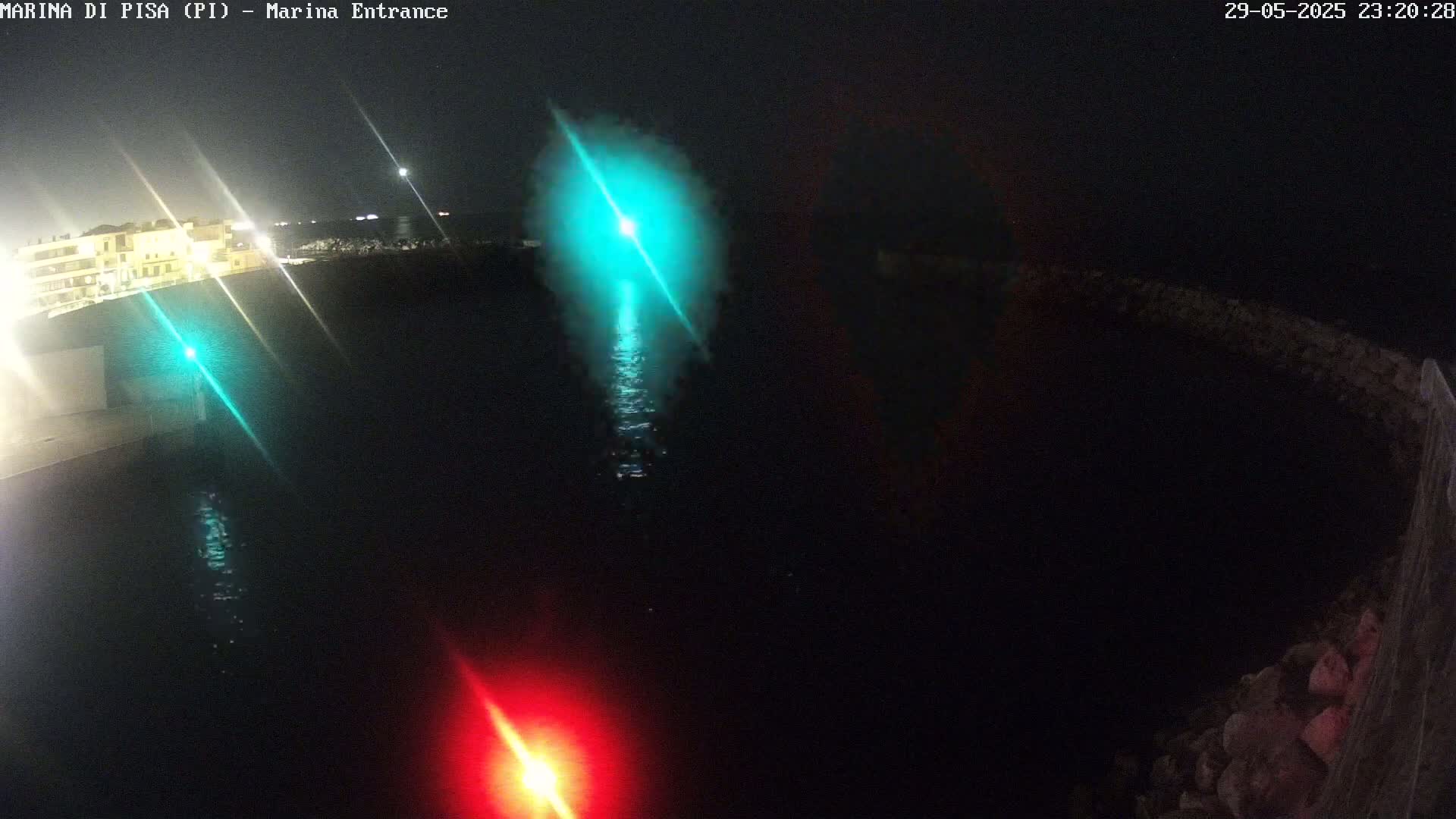 A nighttime view of a dark body of water with a green and a red light reflecting on the surface near a stone wall and buildings in the distance.