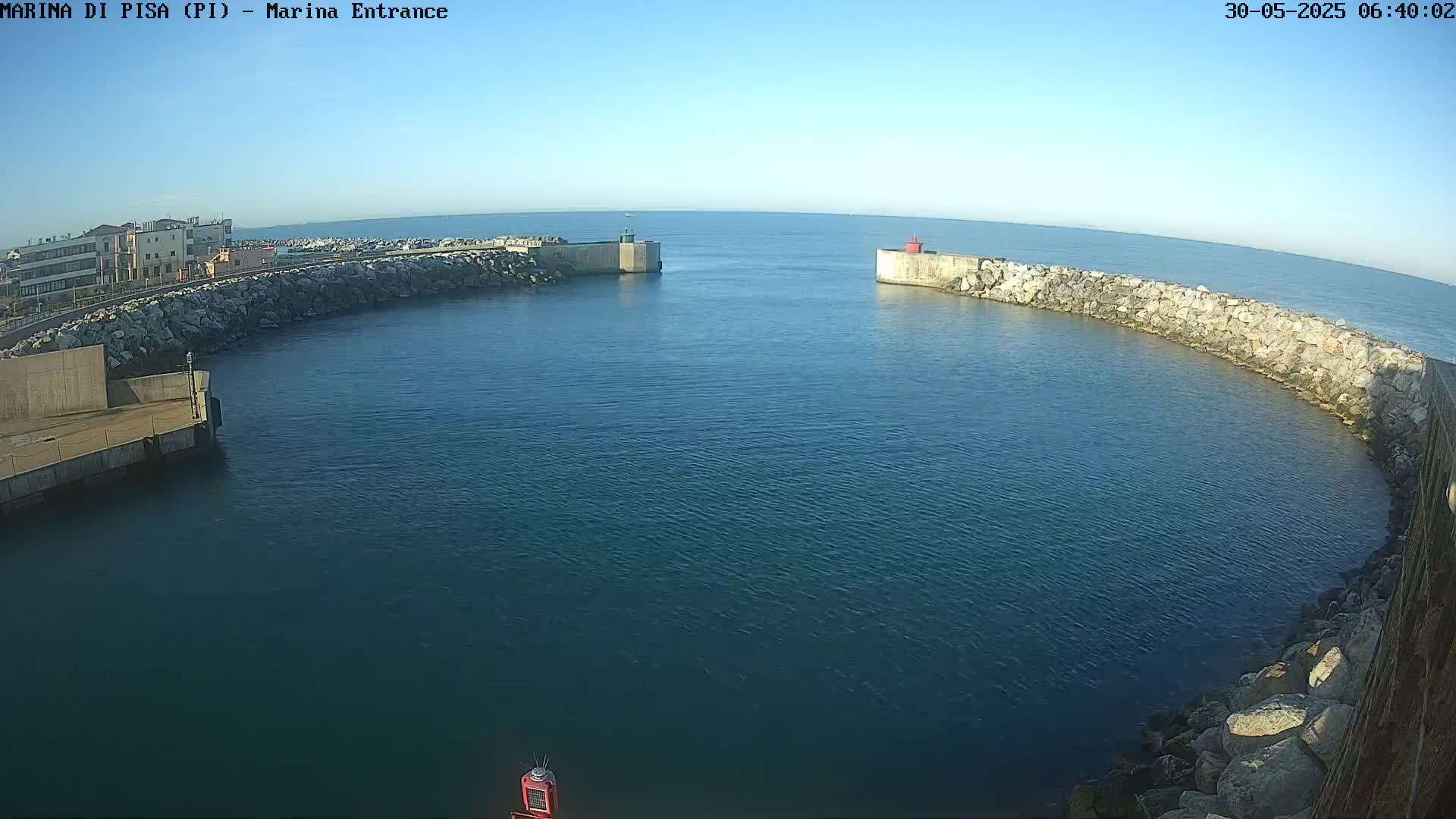 A calm, clear day reveals a marina entrance with stone walls and calm, dark-blue water.