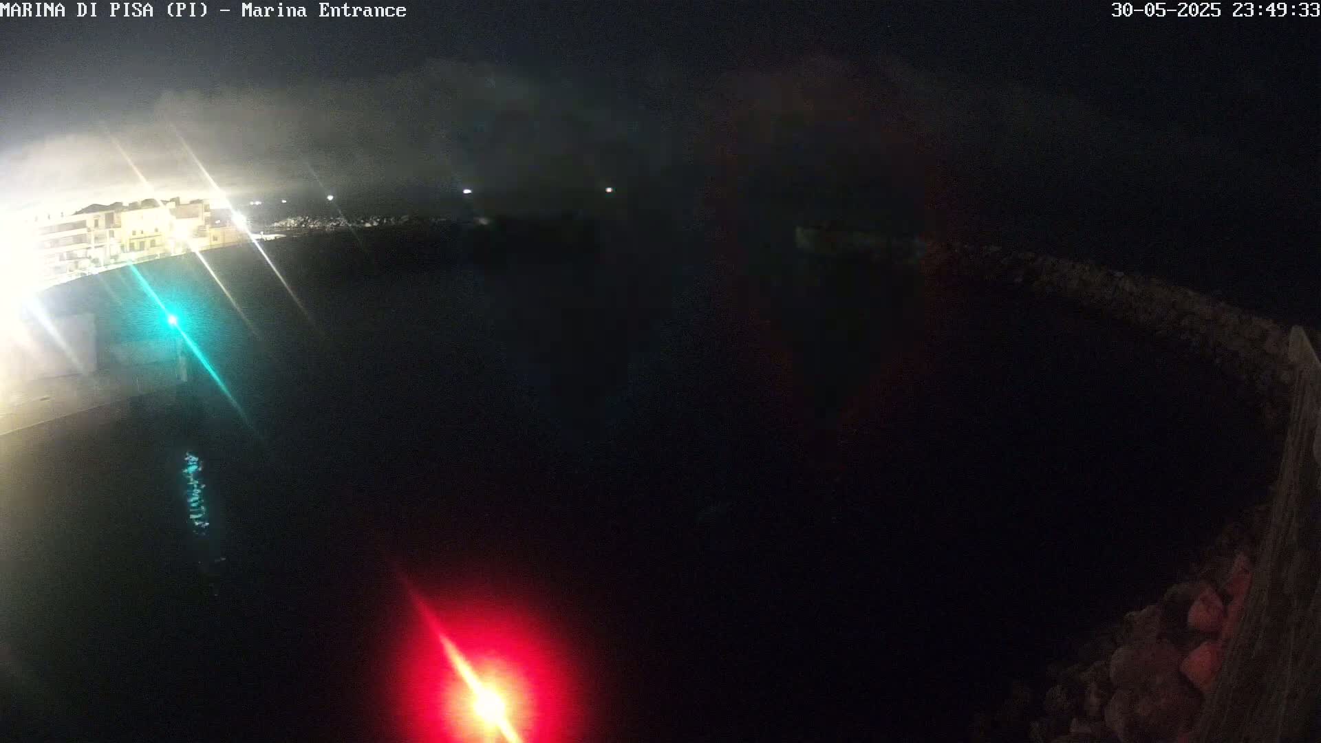 A nighttime, dark view of a harbor entrance with a building visible in the distance and a red and green light visible in the water.