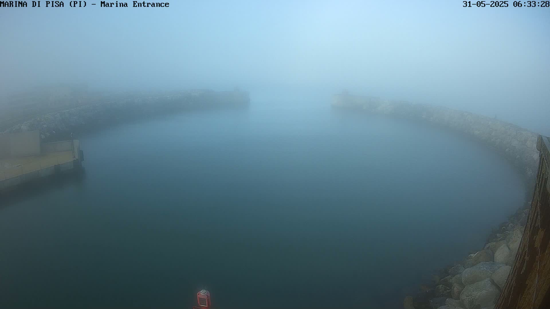 A foggy harbor entrance is seen from an elevated perspective, showing calm water enclosed by stone walls.