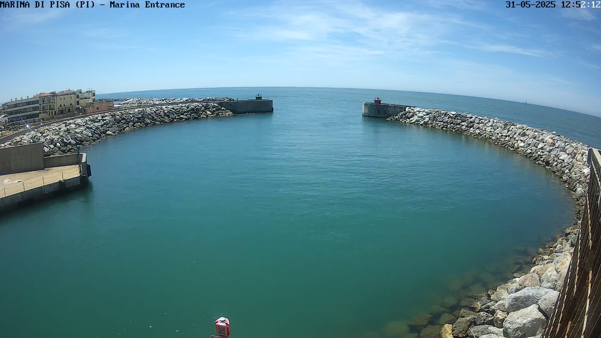 A harbor entrance with teal water under a mostly sunny sky is framed by two concrete walls lined with large rocks.