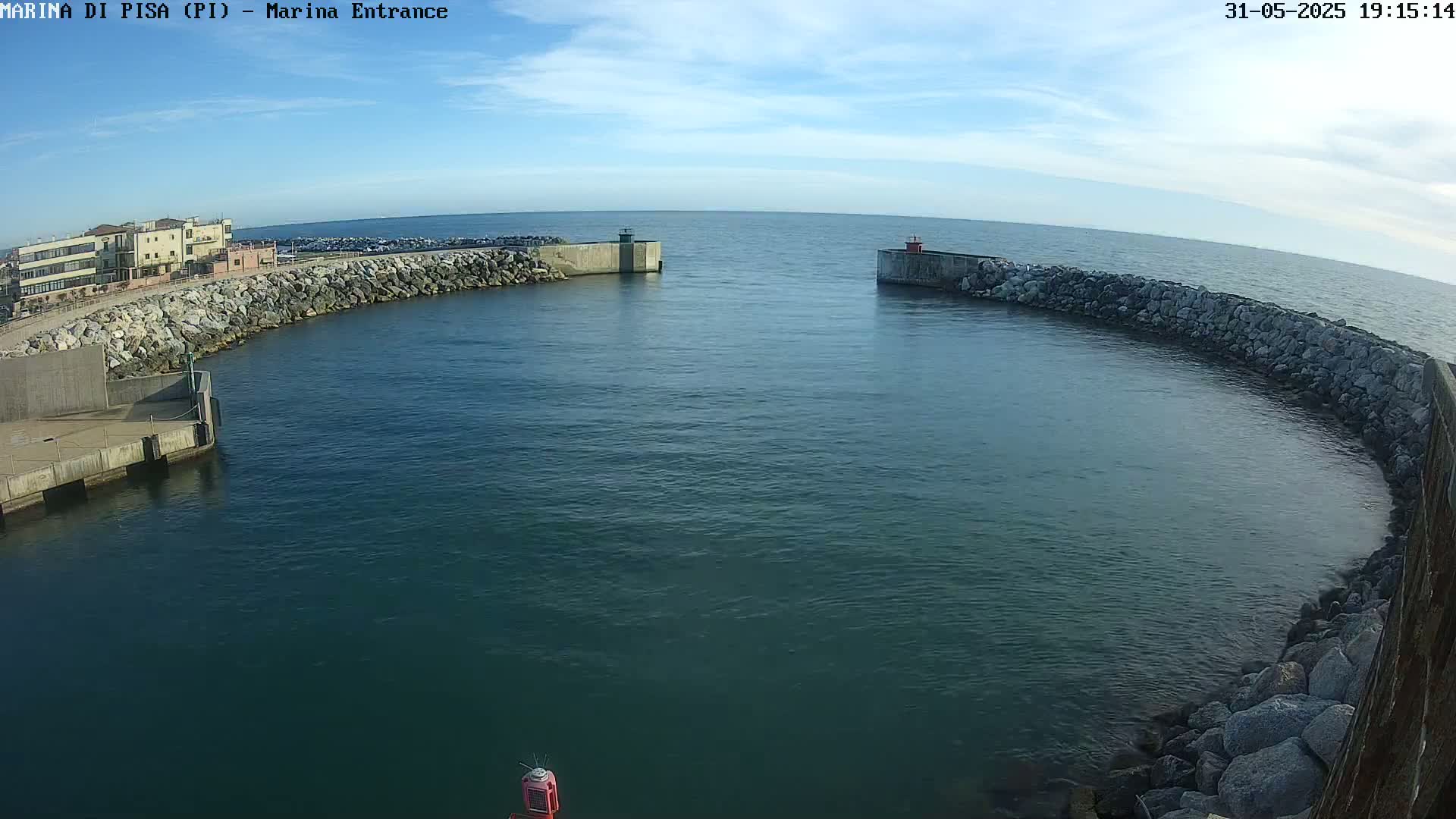 A harbor entrance with calm water under a mostly clear sky, featuring rock walls and a building in the background.
