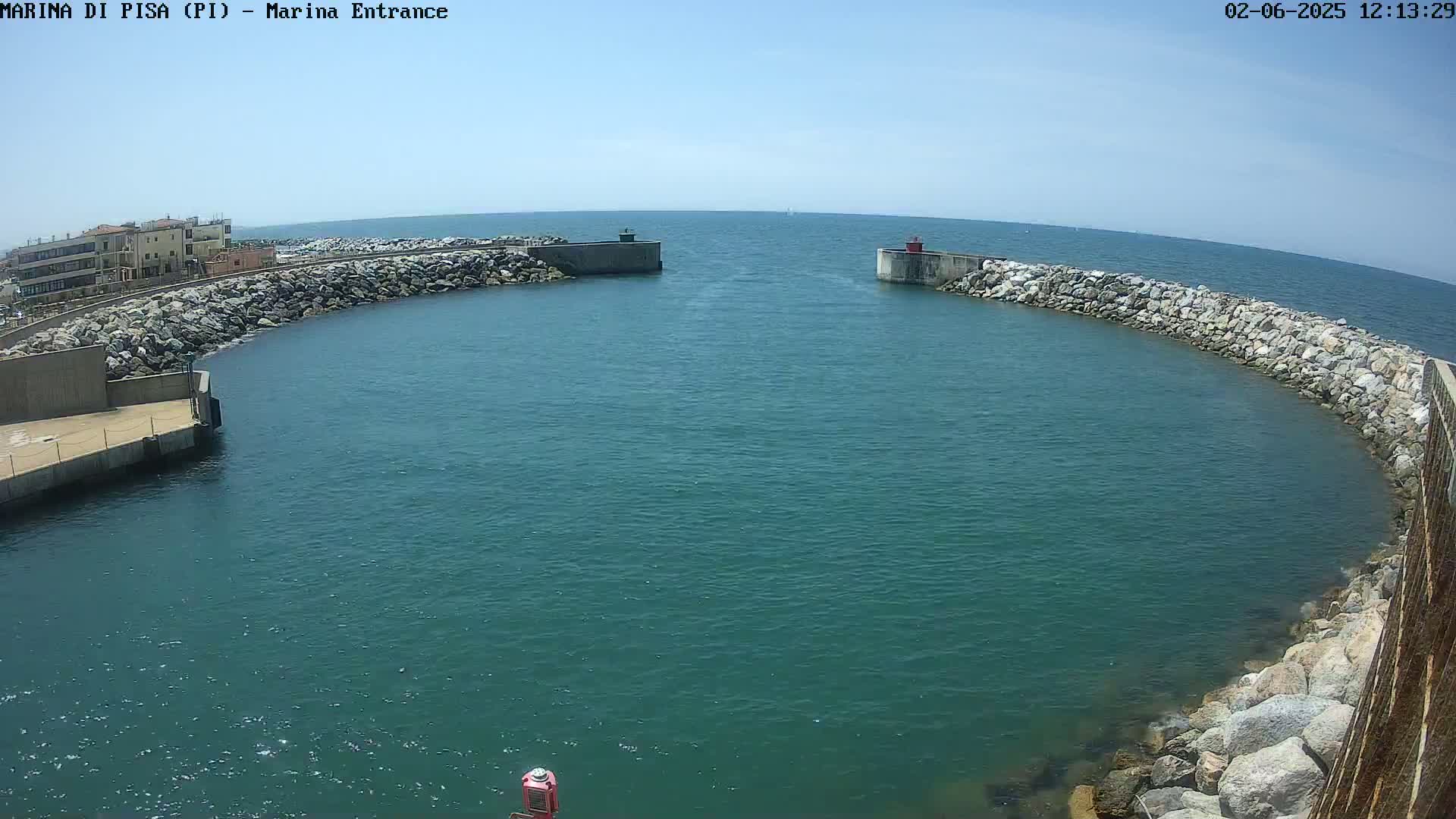 A harbor entrance with calm, teal water is bordered by rock walls and a building in the background under a clear, sunny sky.