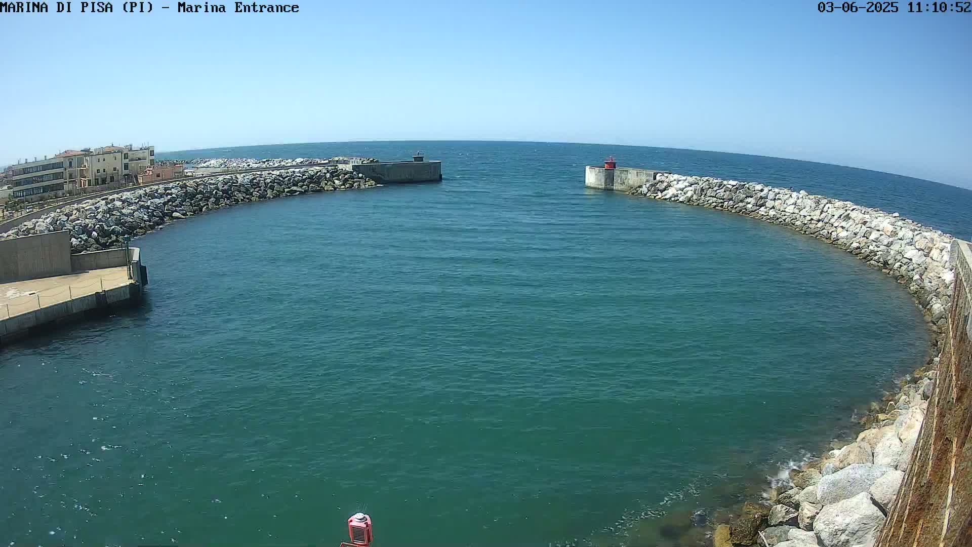 A marina entrance with calm, teal water is framed by rock walls and buildings under a clear, sunny sky.