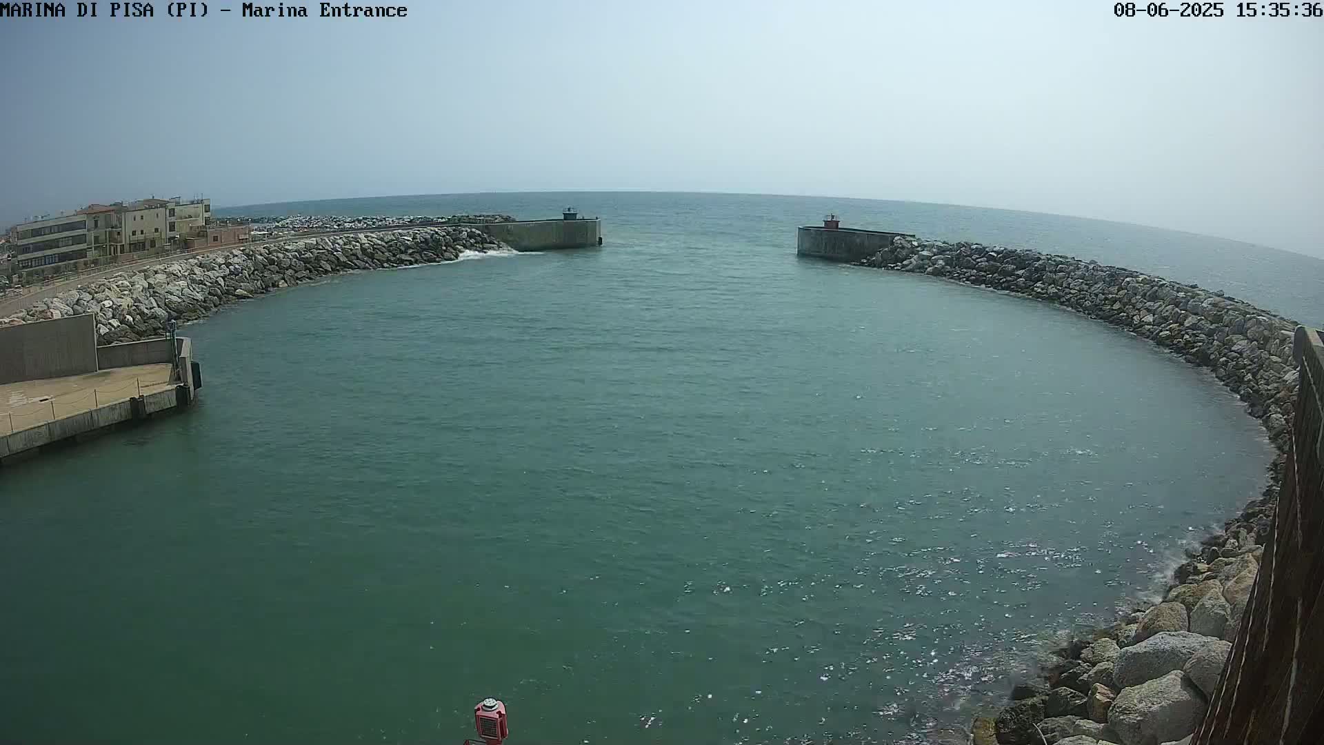 A harbor entrance with calm, greenish-blue water is bordered by rock walls and a building on the left, under a hazy sky.