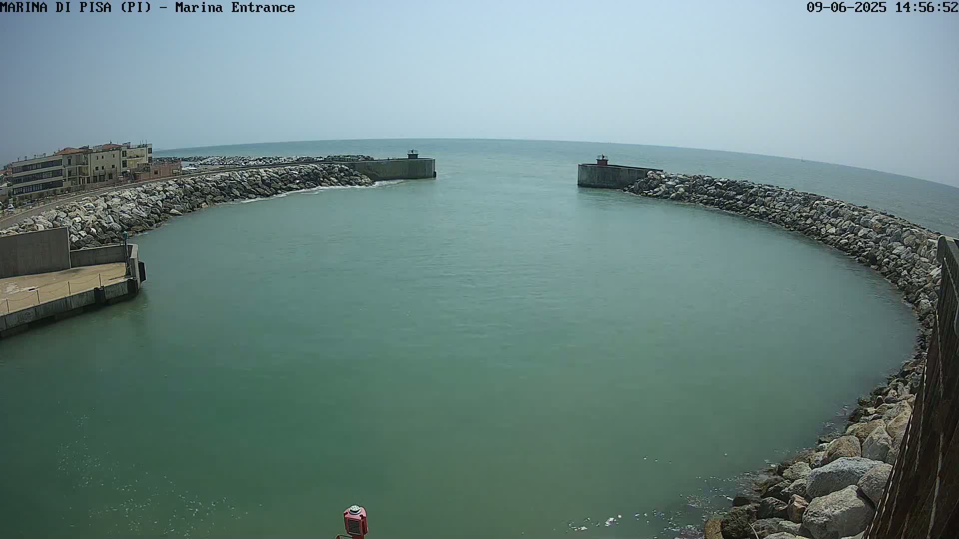 A harbor entrance with calm, greenish water is enclosed by rock walls and a building is visible in the background under a clear, sunny sky.