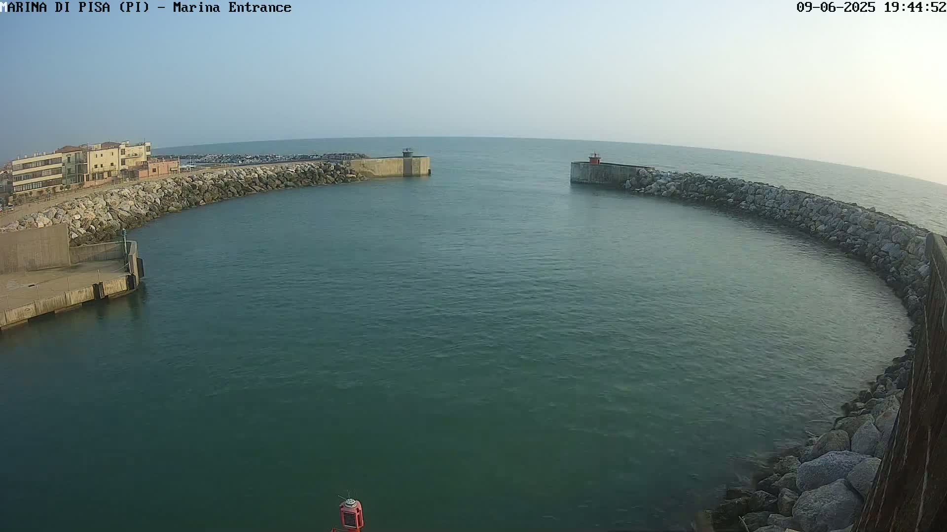 A calm, teal-colored harbor is nestled between two rocky breakwaters on a clear day.