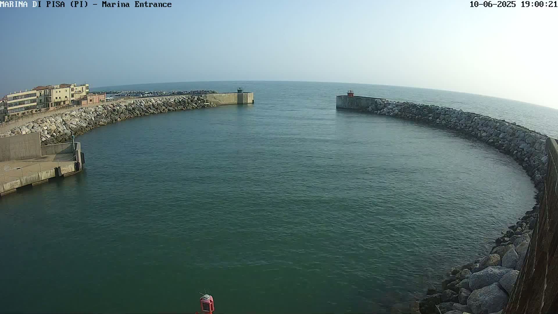 A calm, clear day shows a marina entrance with two stone breakwaters curving into a dark-green body of water.
