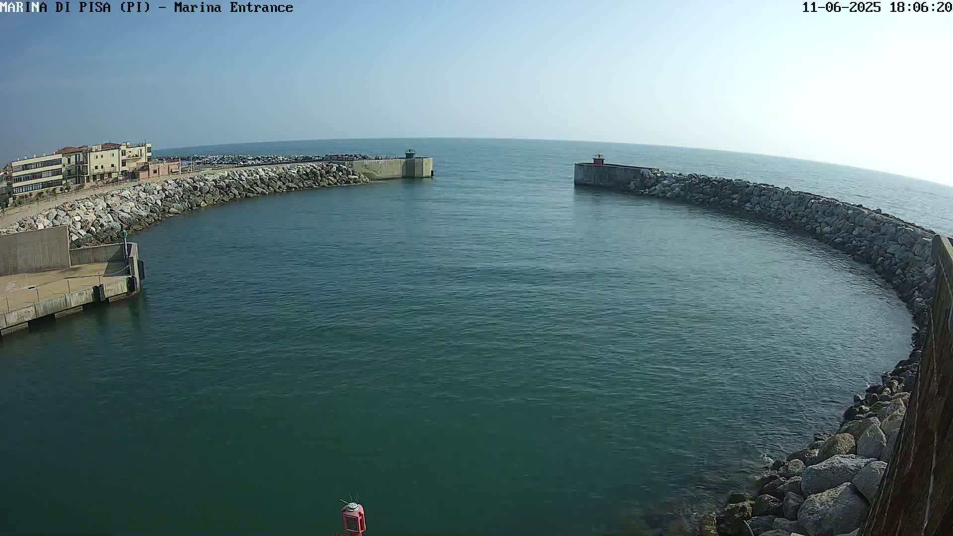 A harbor entrance with calm, clear water is nestled between two stone breakwaters on a sunny day.