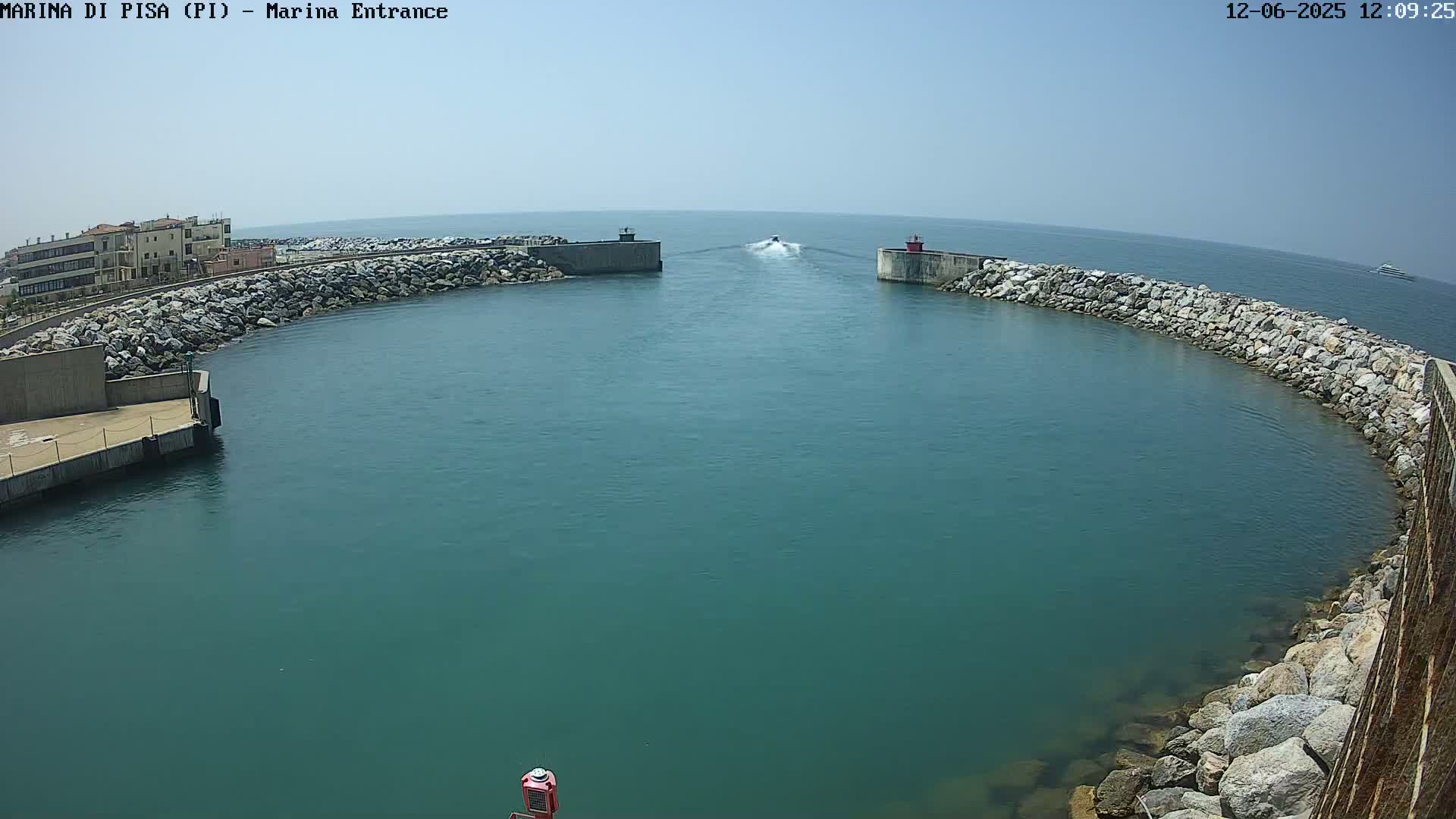 A small motorboat passes through a harbor entrance on a sunny day, with calm teal water and rocky breakwaters.