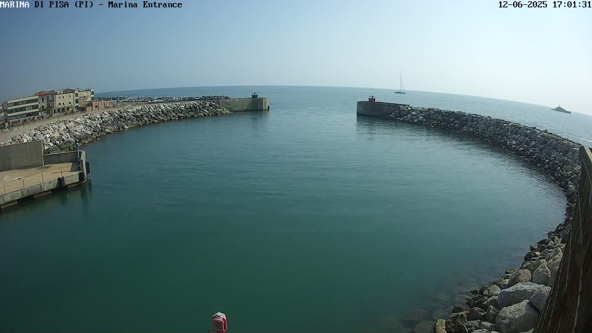 A calm, clear day shows a marina entrance with a curved rock wall, calm teal water, and a few boats in the distance.