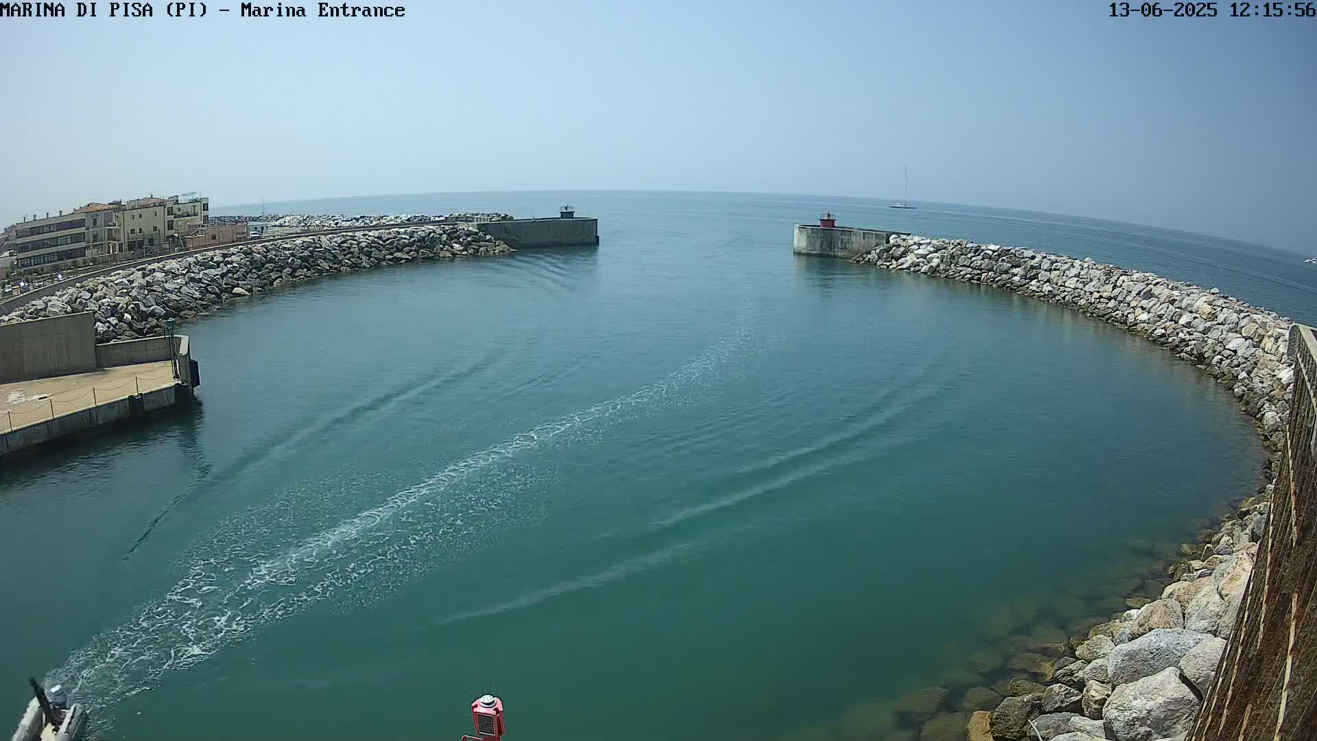 A boat's wake streaks across the teal water of a harbor entrance on a sunny day, bordered by rock walls and buildings in the distance.