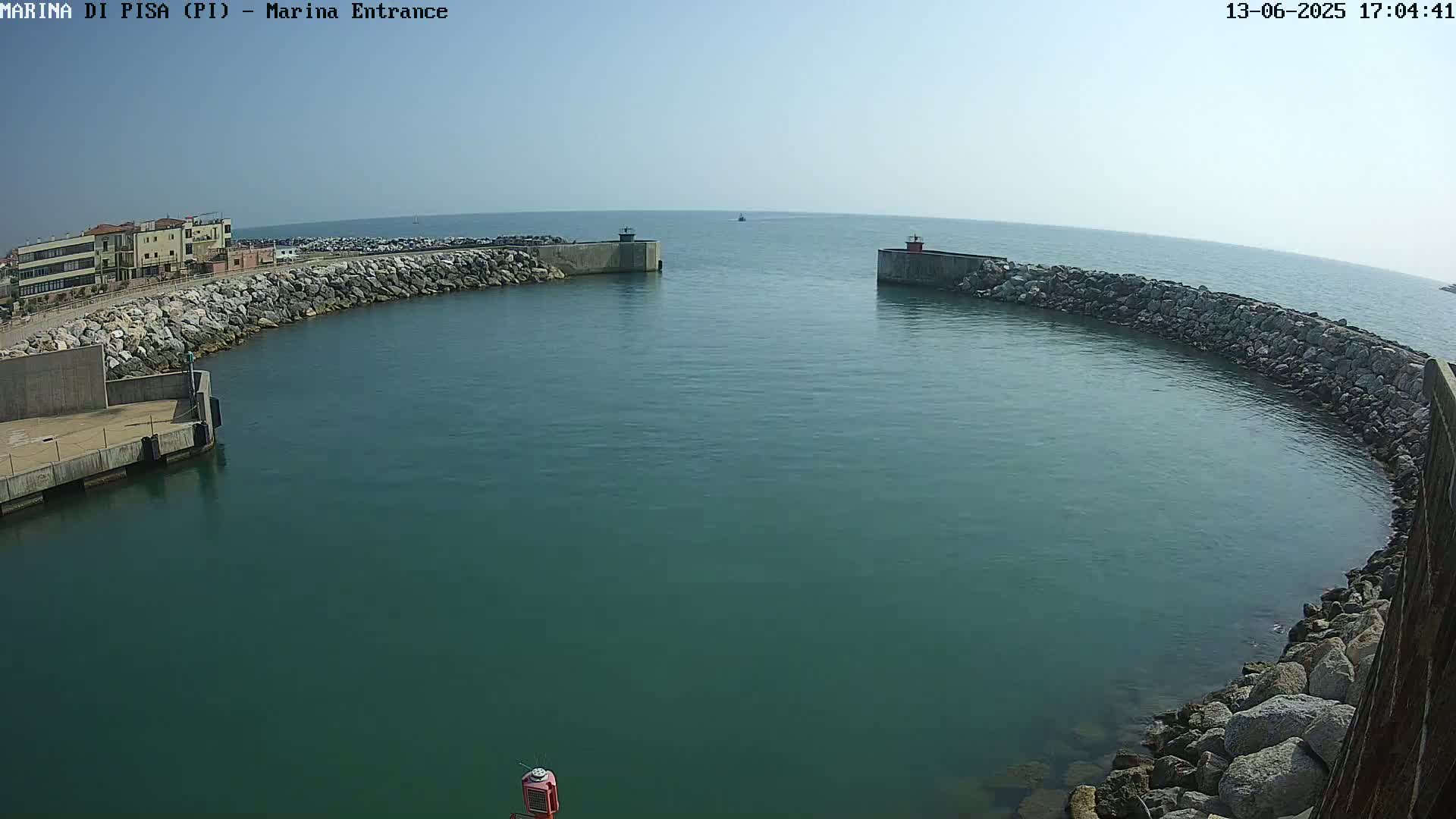 A harbor entrance with calm, teal water is bordered by stone walls, with buildings visible in the background under a clear sky.