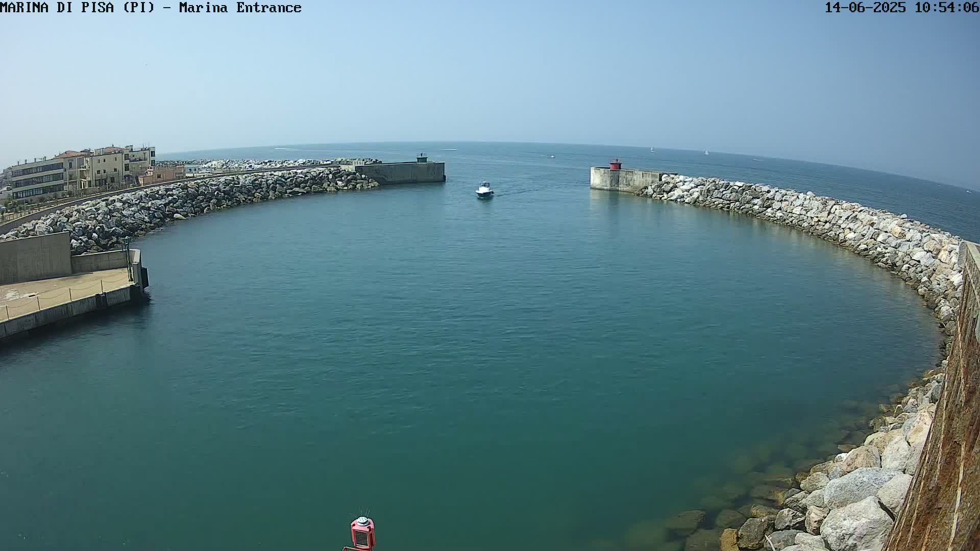 A small motorboat travels through a calm, clear harbor entrance bordered by rock walls and buildings under a bright, sunny sky.