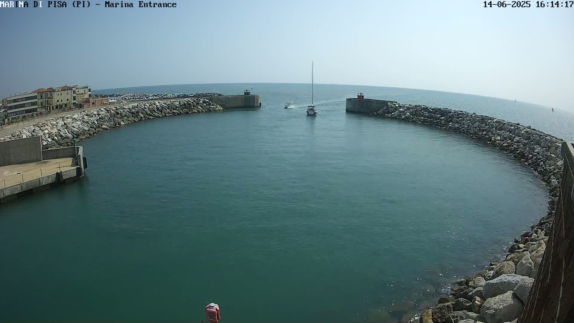 A sailboat and a smaller motorboat navigate the entrance to a harbor on a calm, sunny day, passing between two rock-and-concrete breakwaters.