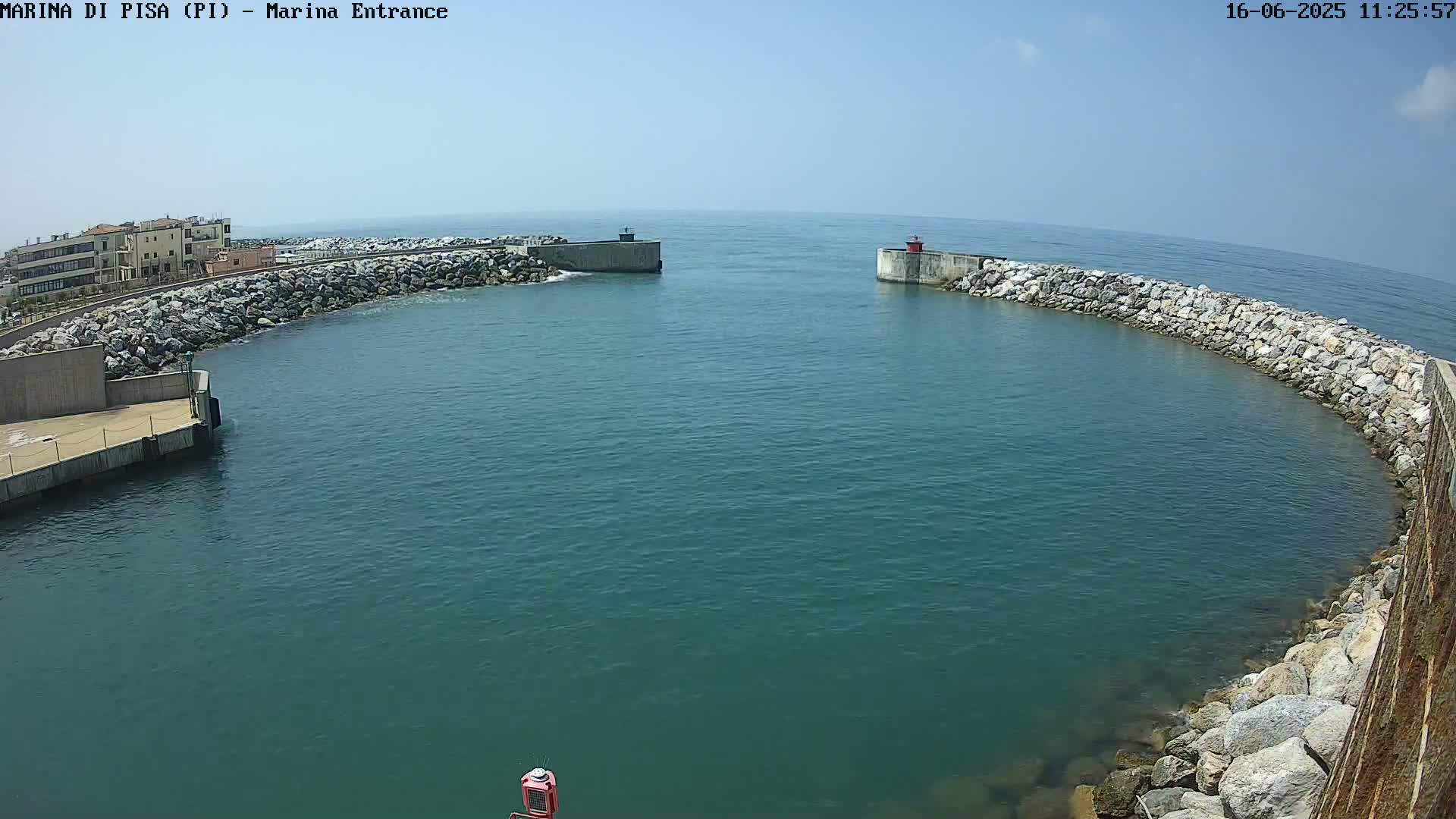 A harbor entrance with calm, blue water is bordered by rock walls and a building in the background under a clear, sunny sky.