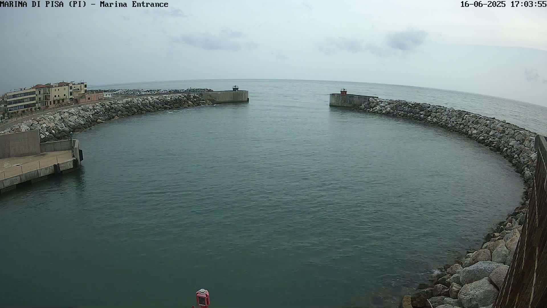 A calm, overcast day reveals a harbor entrance with stone walls, buildings on one side, and a mostly calm body of water extending to a horizon.