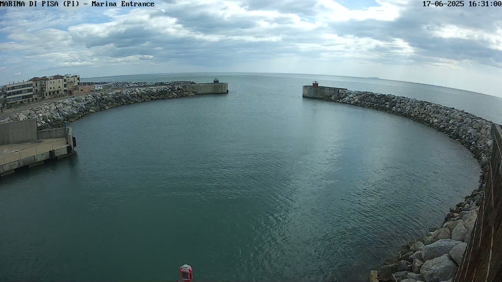 A partly cloudy day shows a marina entrance with calm, dark-green water between two rock-lined walls.