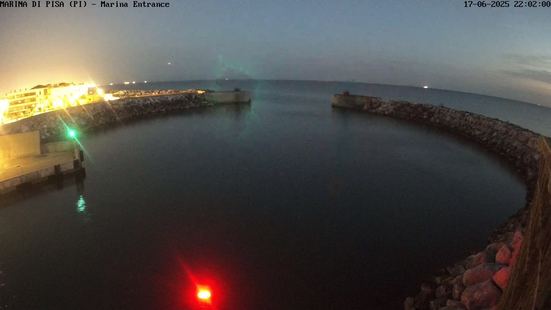 A dark, calm harbor at night is enclosed by a low stone wall with a building lit up on the left, under a twilight sky.