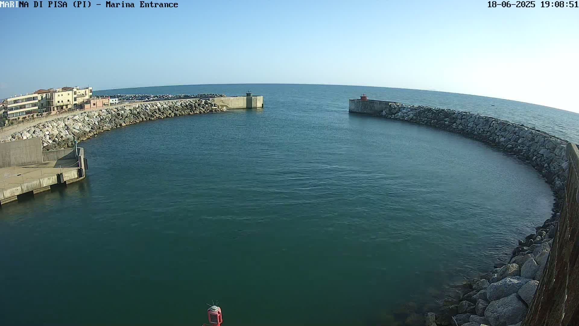 A calm, clear day shows a harbor entrance with a stone breakwater curving around a protected body of dark-green water, buildings visible in the background.