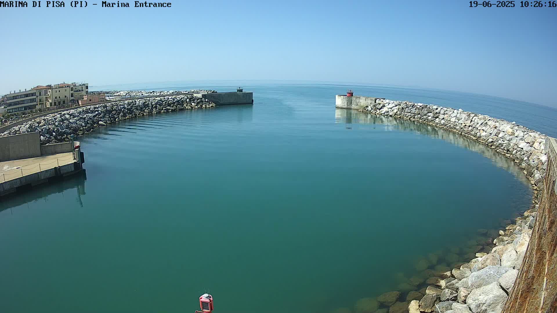 A harbor entrance with calm, teal-colored water under a clear blue sky is bordered by rock walls and a building in the distance.