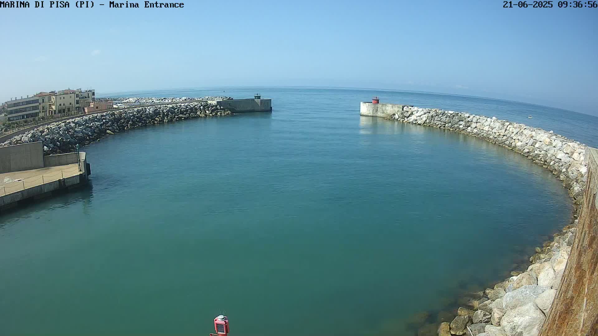 A calm, clear day shows a harbor entrance with teal water enclosed by rock and concrete walls, extending into a larger body of water.