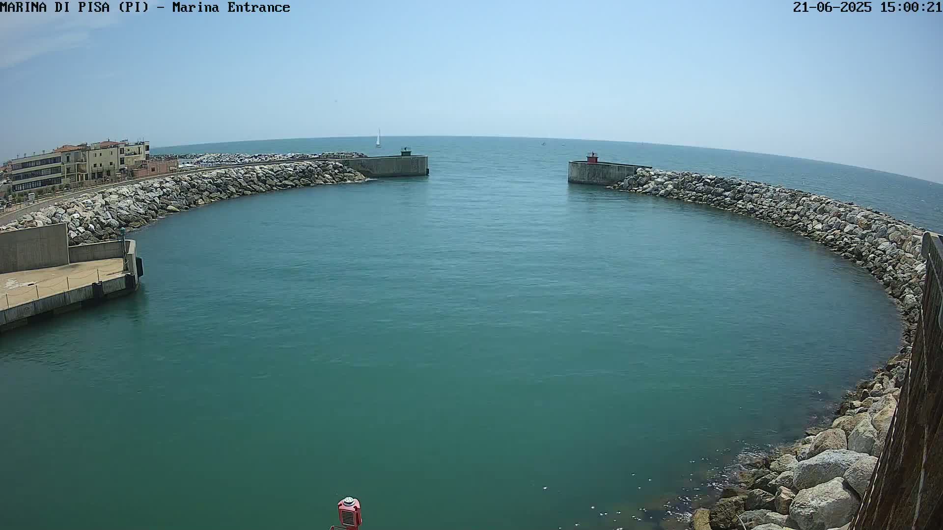 A marina entrance with calm, teal-colored water under a clear blue sky is surrounded by rock walls and a building in the background.