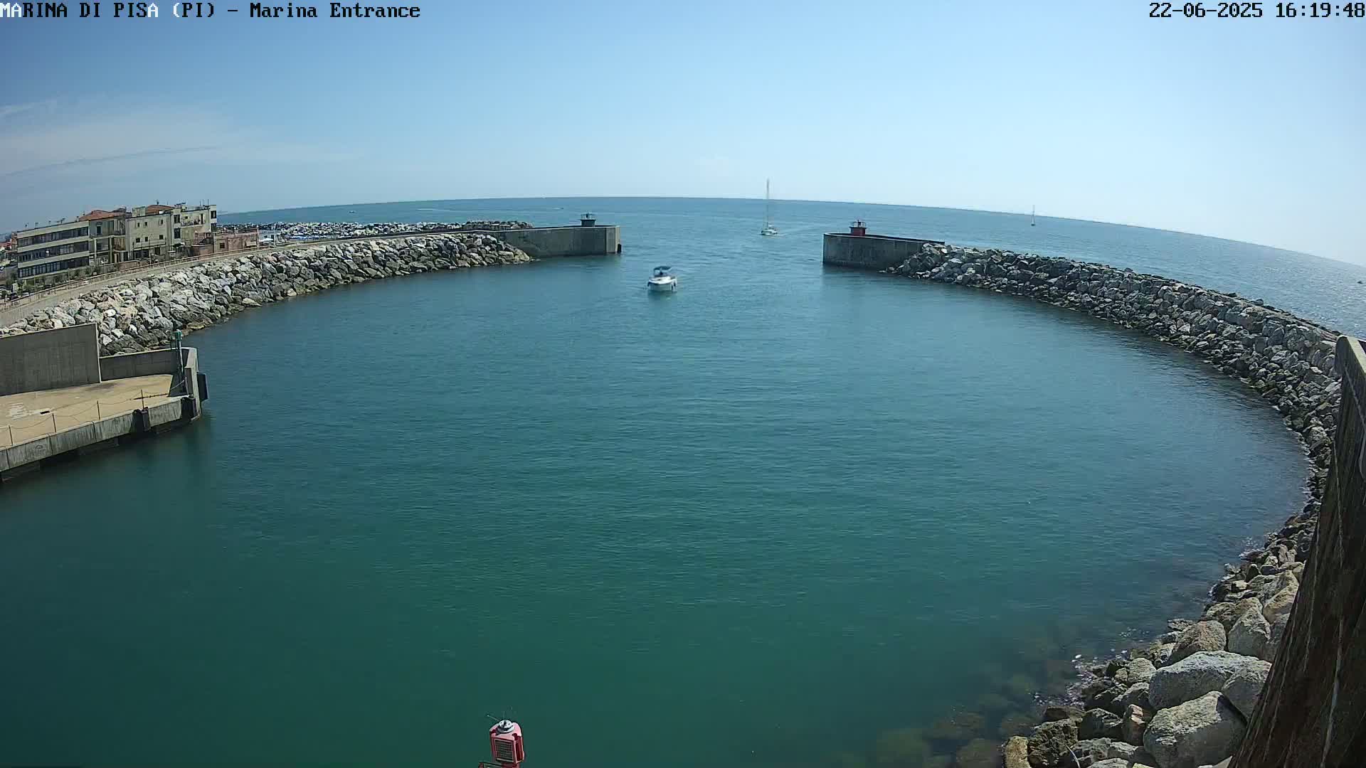 A marina entrance with calm, clear blue water is enclosed by rock walls, with a small boat visible in the channel and buildings in the background under a clear blue sky.