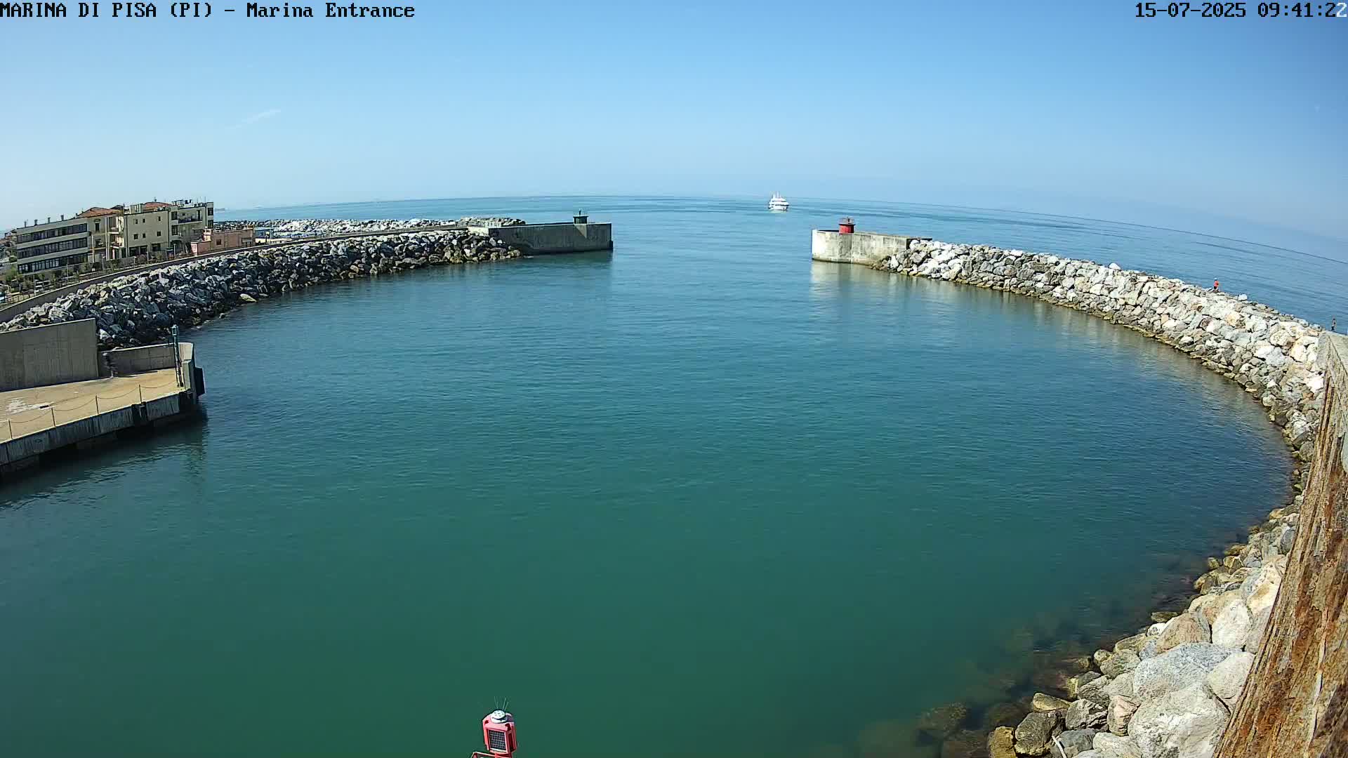 A harbor entrance with calm, teal water is framed by rock walls, with a boat visible in the distance under a clear blue sky.