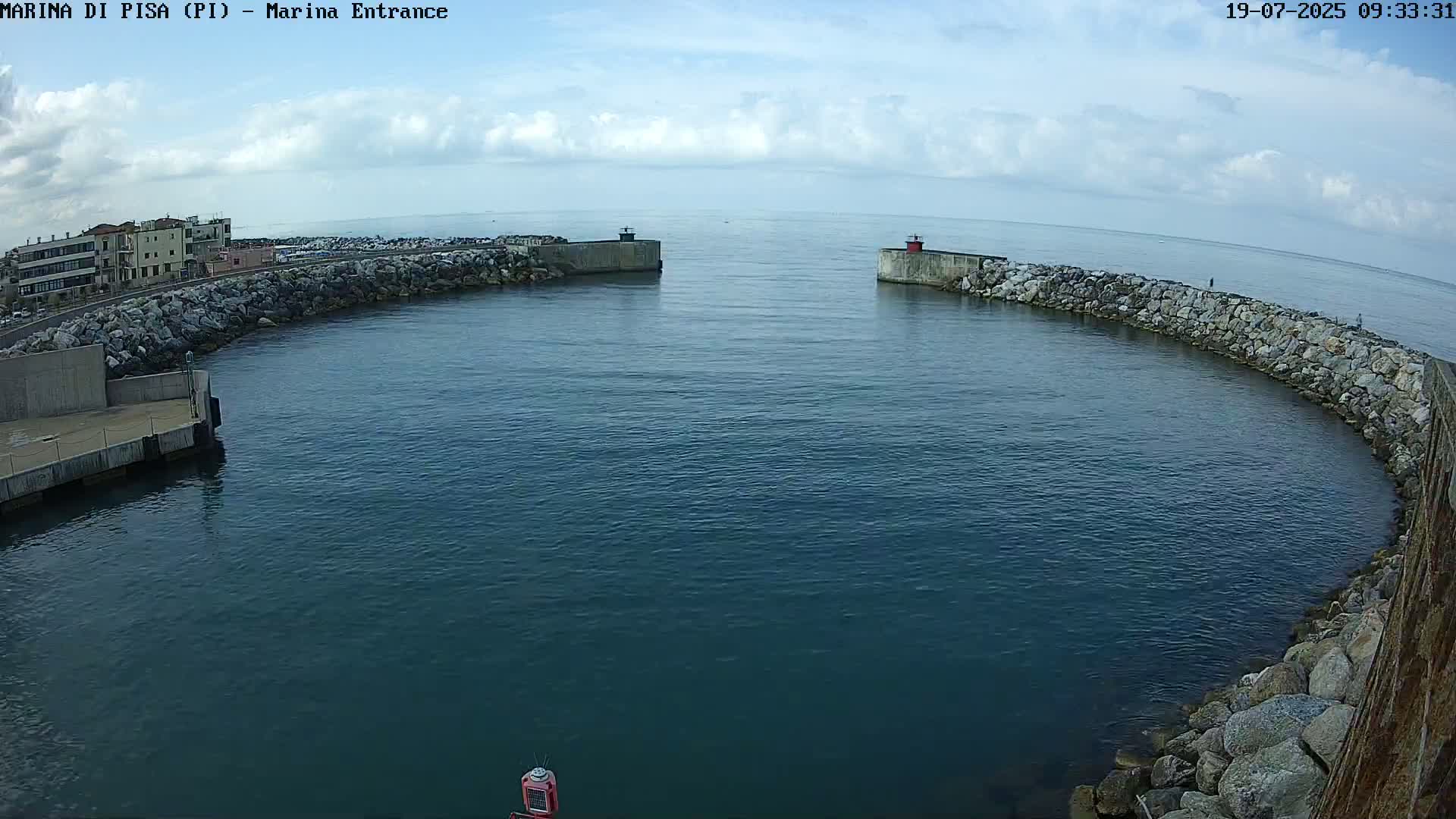 A partly cloudy day shows a harbor entrance with calm, dark-blue water between two stone walls, and a small town in the background.