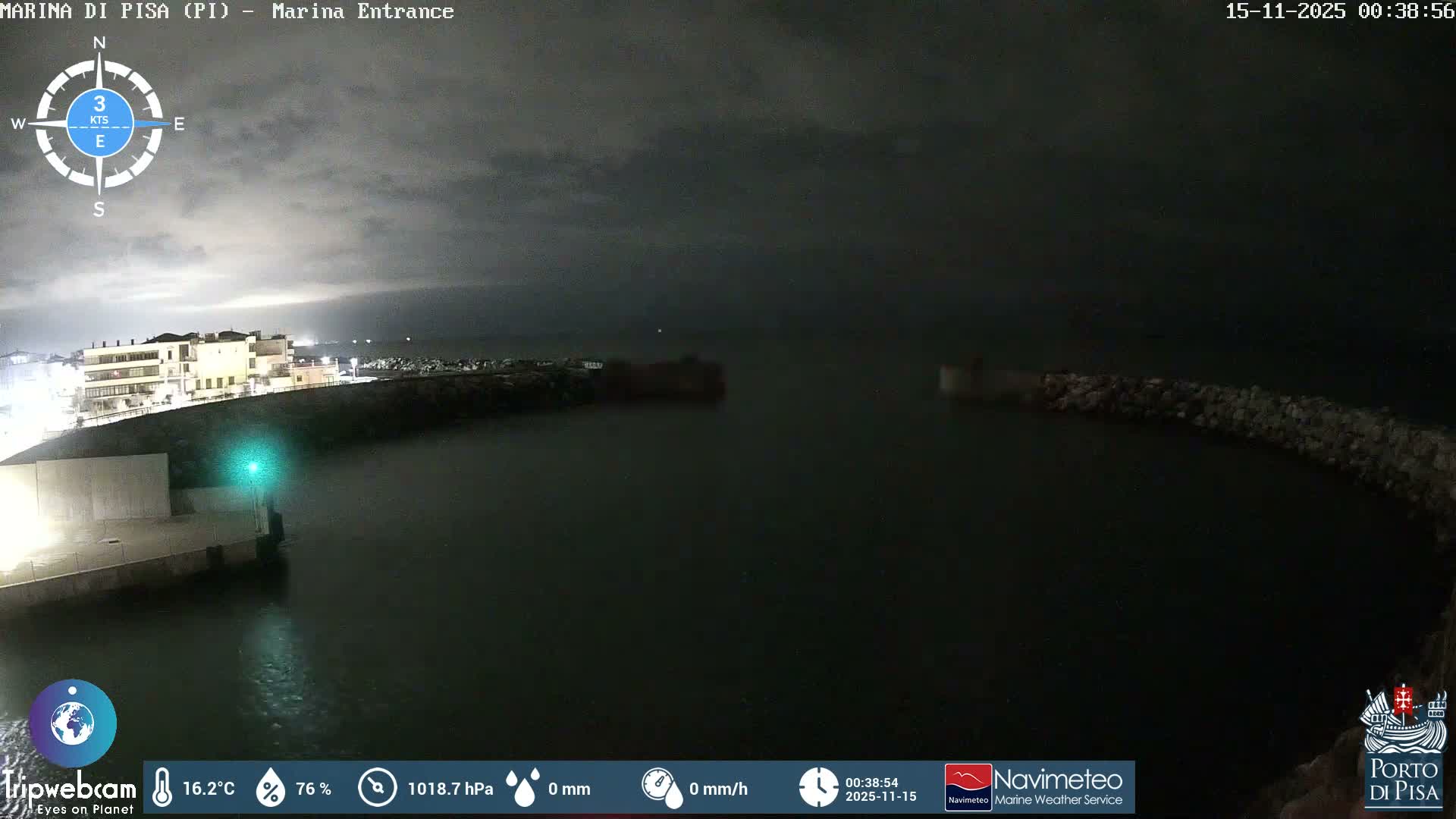 A night view reveals a marina entrance with illuminated buildings and a green navigation light on the left, breakwaters extending into calm water, all under a heavily overcast sky.