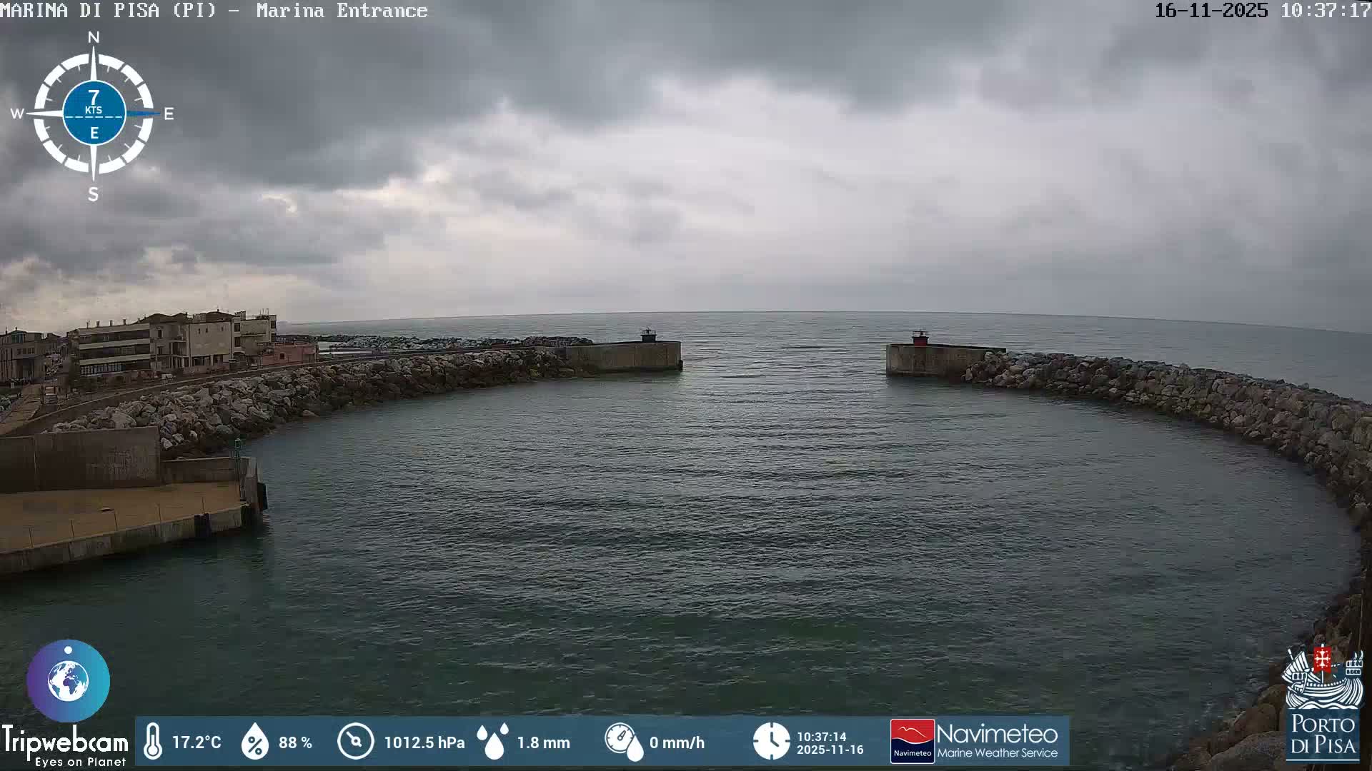 The image shows a marina entrance with two rock jetties, each capped by a small lighthouse, opening to the sea under a heavily overcast sky, with buildings lining the left shore.