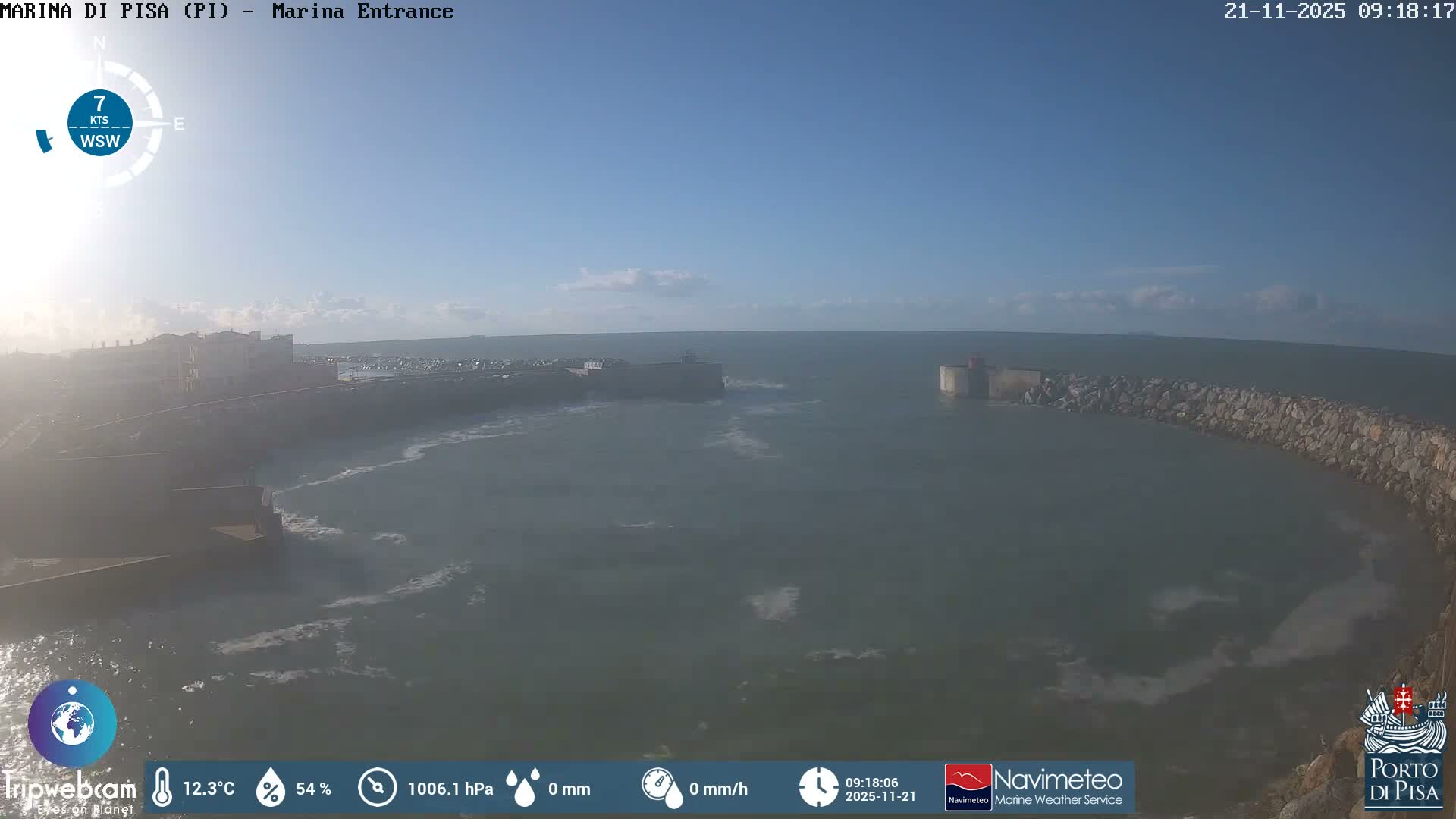 The image shows a sunny marina entrance with choppy green-blue water and rocky breakwaters under a mostly clear blue sky with a few wispy clouds on the horizon, bordered by distant buildings on the left.
