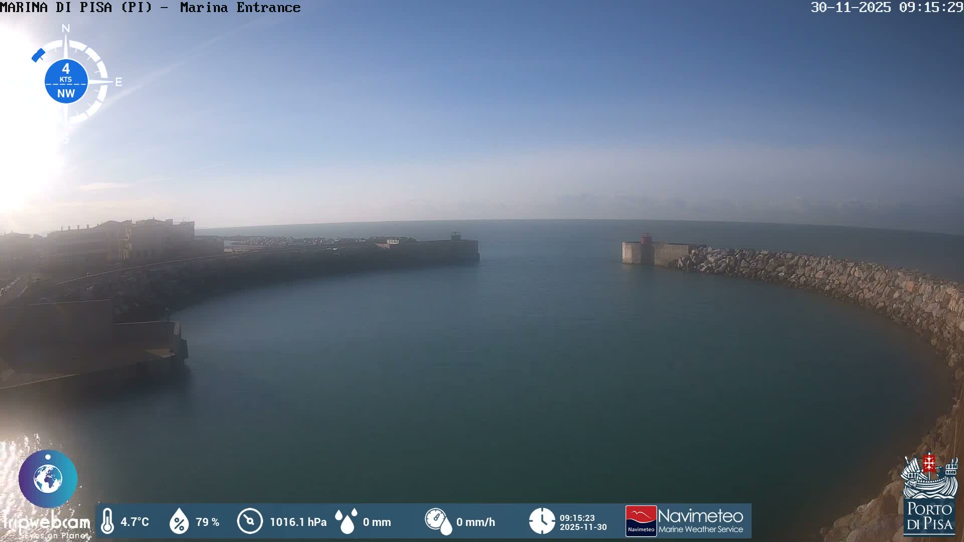 A clear, sunny day reveals the calm, teal waters of a marina entrance, flanked by urban buildings on the left and a long rocky breakwater with a red lighthouse beacon on the right, leading out to a hazy horizon.