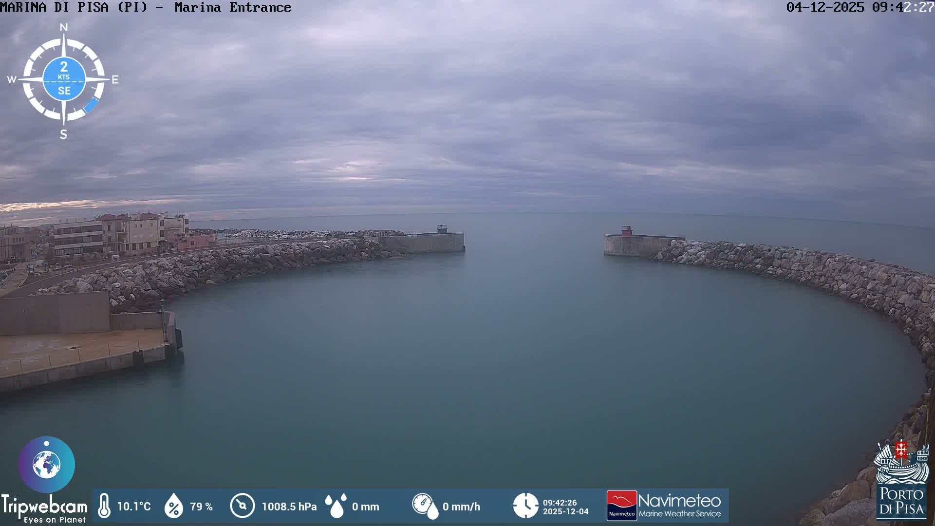 A calm marina entrance with two small lighthouses marking the gap between rocky breakwaters is viewed under a heavily overcast and dull grey sky, with buildings visible along the distant left bank.
