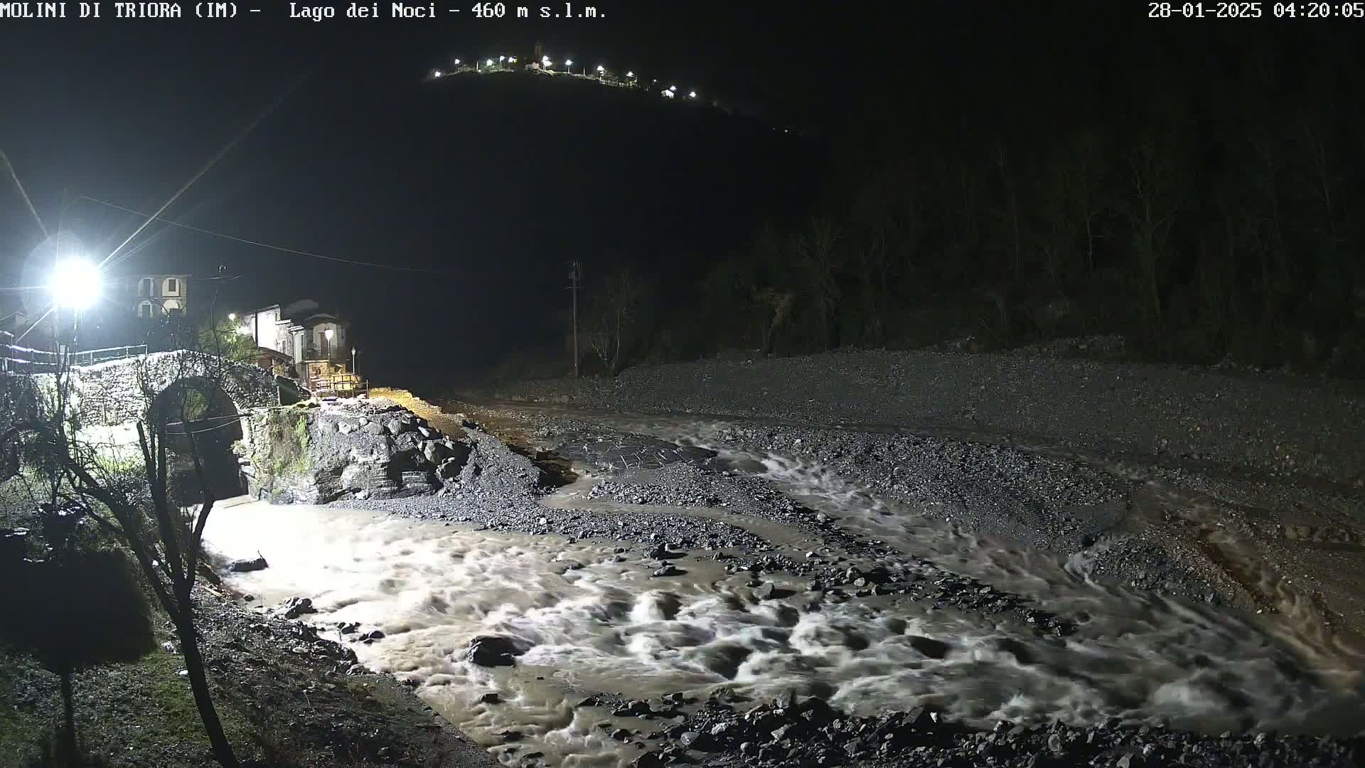 A fast-flowing, muddy river at night rushes under a stone bridge near a small village, illuminated by artificial lights.
