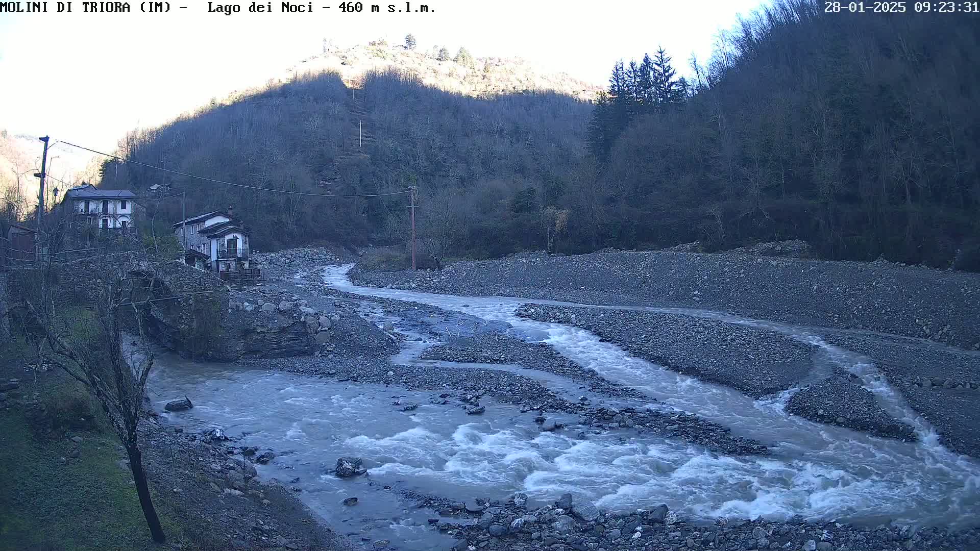 A swiftly flowing river snakes through a rocky riverbed, near a small village nestled against a hillside under a mostly clear sky.