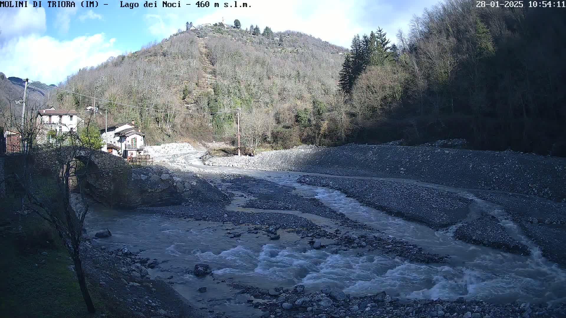 A swiftly flowing river cuts through a rocky riverbed beneath a partly sunny sky, passing by a small village nestled against a hillside of leafless trees.
