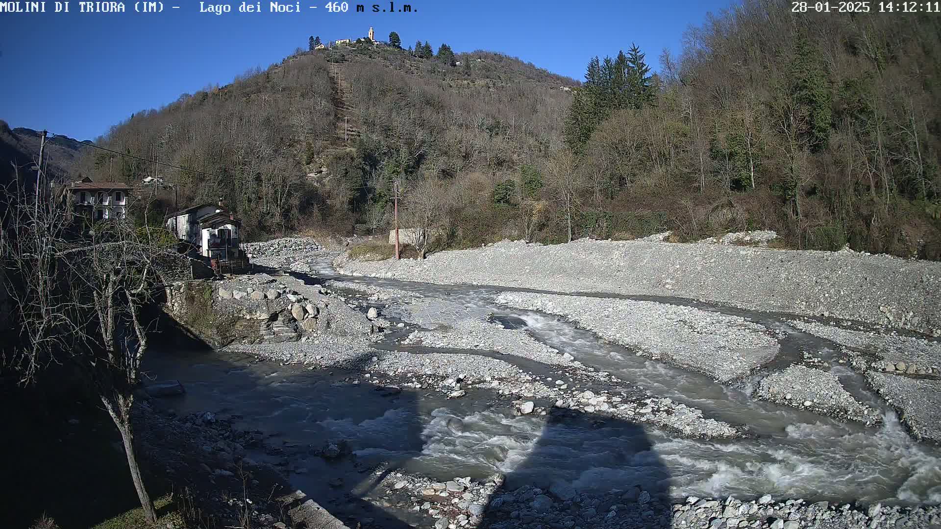 A rocky river flows through a valley under a clear blue sky, past buildings and leafless trees on a hillside.