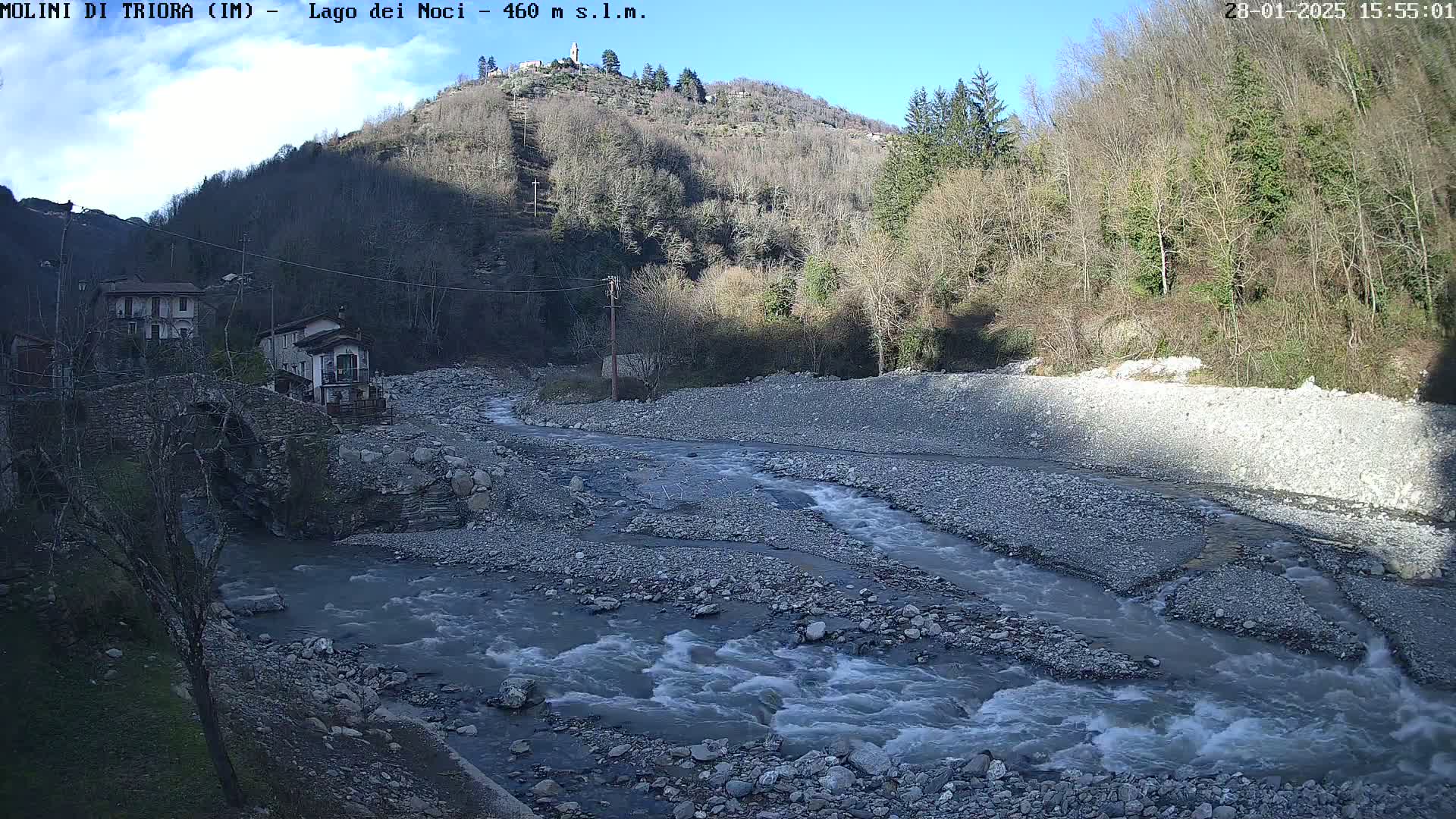 A rocky river flows through a valley under a mostly sunny sky, past a stone bridge and houses nestled against a hillside covered in leafless trees.