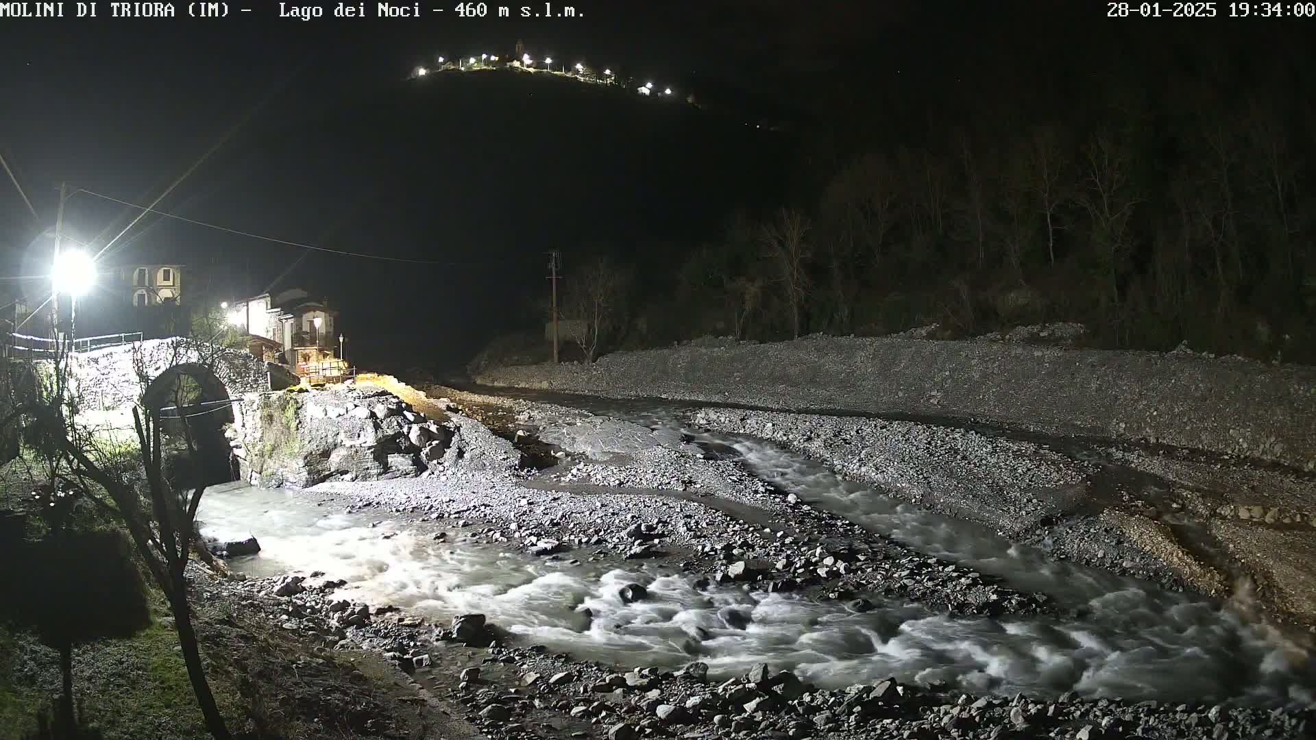 A swiftly flowing river at night, illuminated by artificial light, runs past a stone bridge and a small village nestled at the foot of a hill.