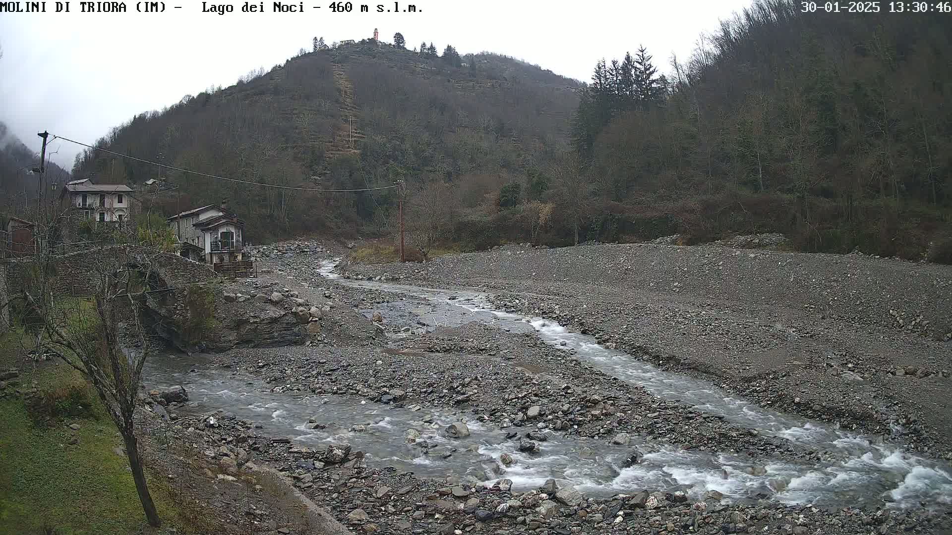 A rocky stream flows through a gray-colored, sparsely vegetated valley under an overcast sky, past a small village nestled against a hillside.