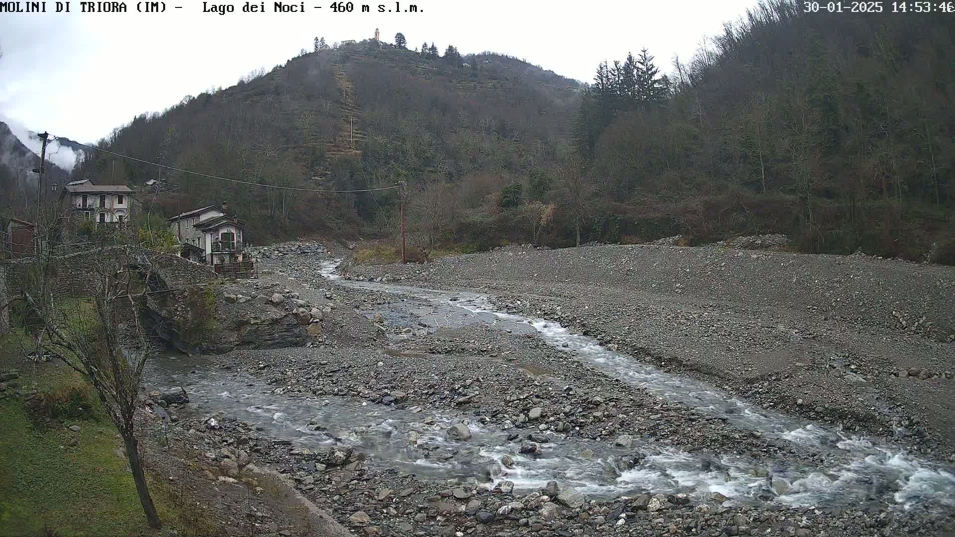 A rocky, shallow river flows through a sparsely vegetated valley under an overcast sky, past a small village nestled against a hillside.