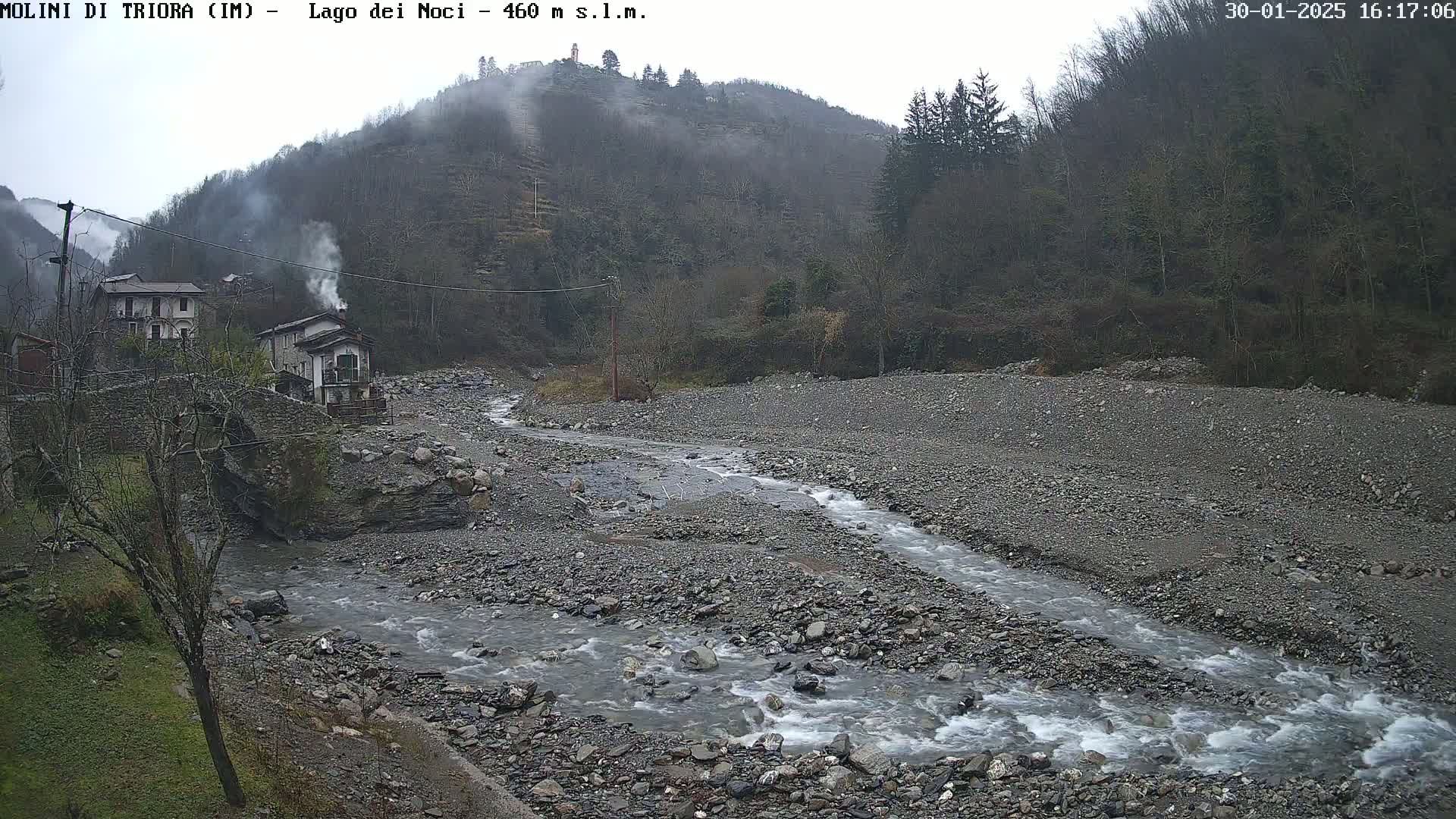 A rocky stream flows through a valley, past a small village nestled against a fog-shrouded hillside under an overcast sky.