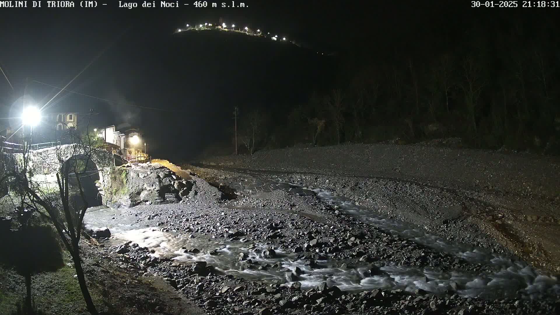 A rocky stream flows through a village at night, illuminated by artificial lights, under a clear, dark sky.