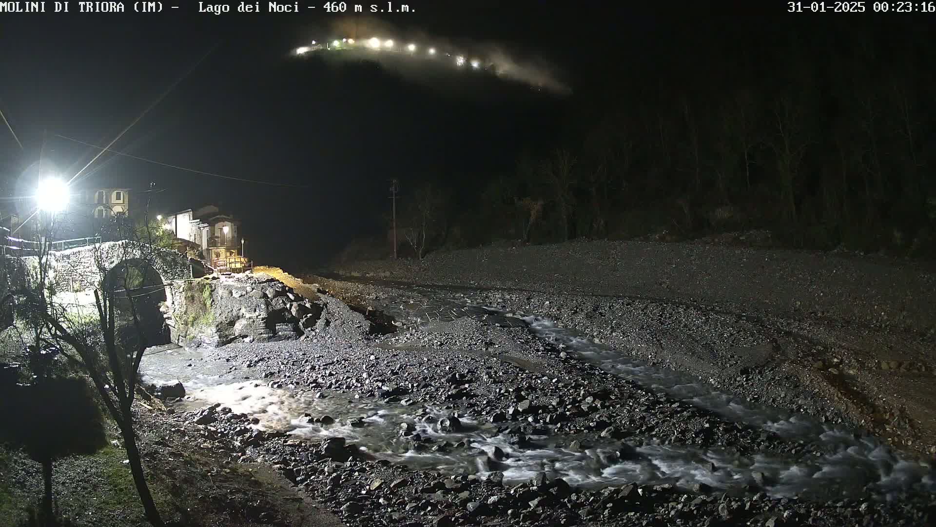 A rocky stream flows at night past a stone bridge and small village under artificial lights, with a hillside visible in the background.