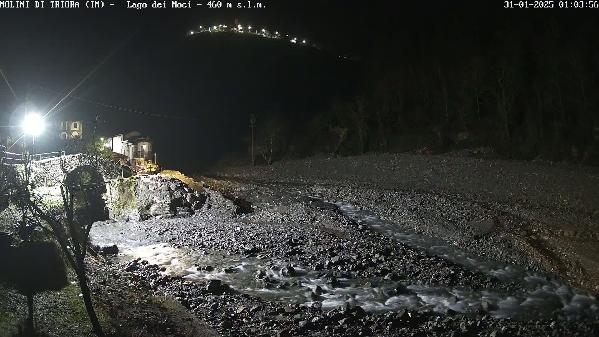 A rocky stream flows under a stone arch bridge at night, illuminated by artificial lights, near a village nestled at the base of a hill.