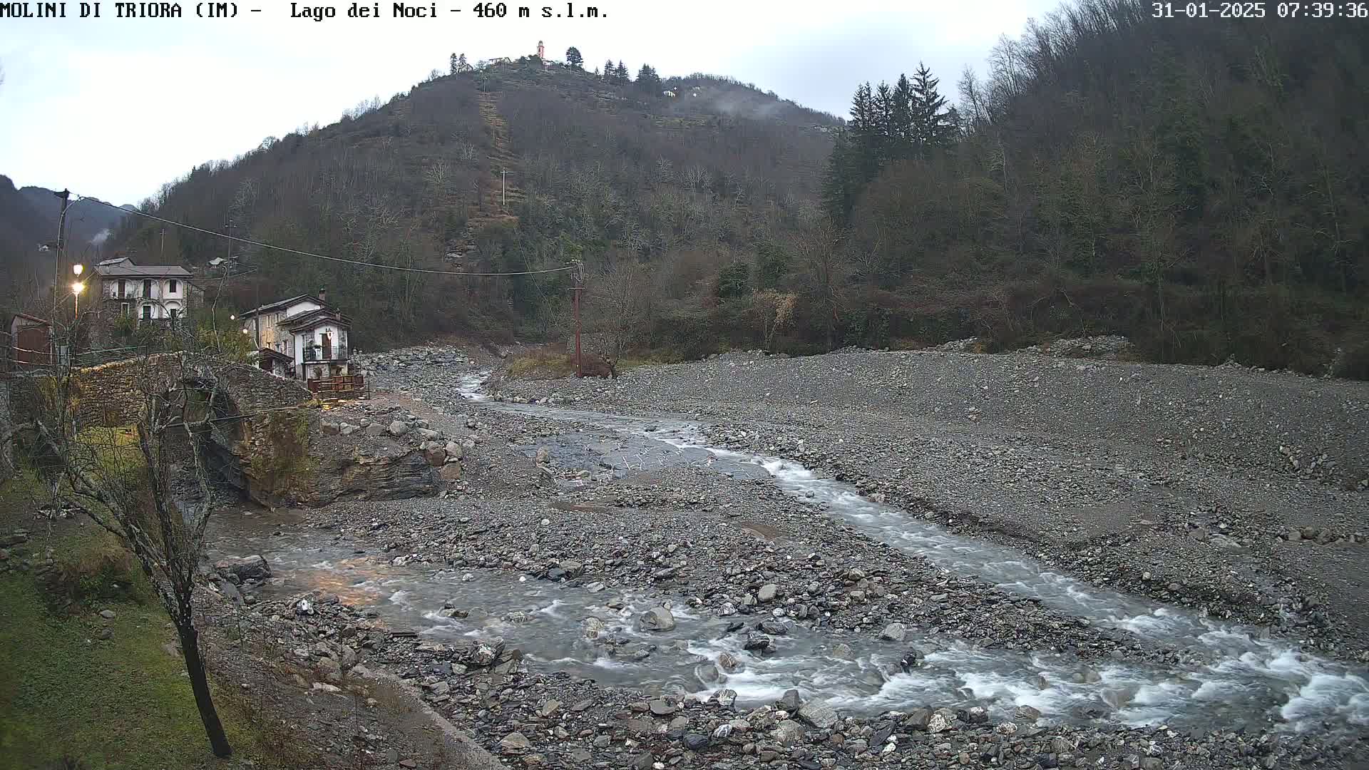 A rocky stream flows through a mostly dry riverbed near a stone bridge and a couple of buildings nestled against a wooded hillside under an overcast sky.