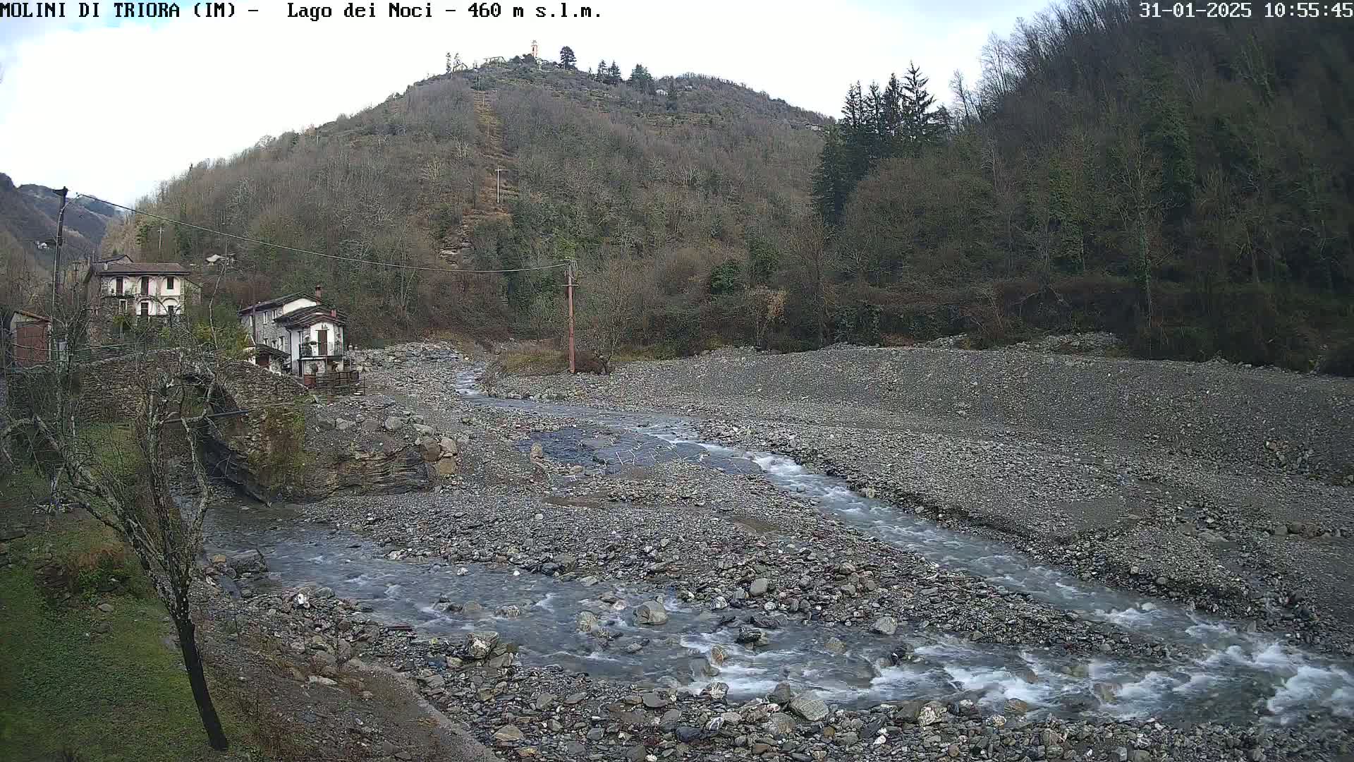 A rocky, grey riverbed with a flowing stream winds through it, past a small village nestled against a hillside under an overcast sky.