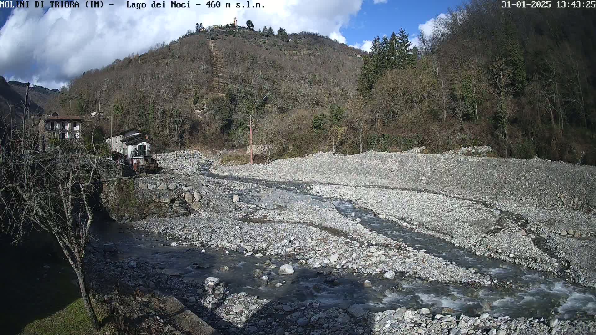 A rocky riverbed with a shallow, flowing stream winds through it, nestled in a valley beneath a partly sunny sky and barren trees on a hillside.